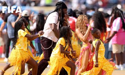 Shakira in a pink and yellow fringed outfit sings into a pink microphone alongside Burna Boy and Uganda's Ghetto Kids dancers on the pitch.