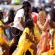 Shakira in a pink and yellow fringed outfit sings into a pink microphone alongside Burna Boy and Uganda's Ghetto Kids dancers on the pitch.