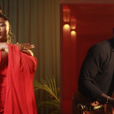 Split-screen music video still from Don't Be Shy featuring Yemi Alade dancing in a sheer red asymmetrical chiffon cape top next to Bien wearing a brown shirt, patterned gold tie, and holding a camera.
