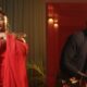 Split-screen music video still from Don't Be Shy featuring Yemi Alade dancing in a sheer red asymmetrical chiffon cape top next to Bien wearing a brown shirt, patterned gold tie, and holding a camera.