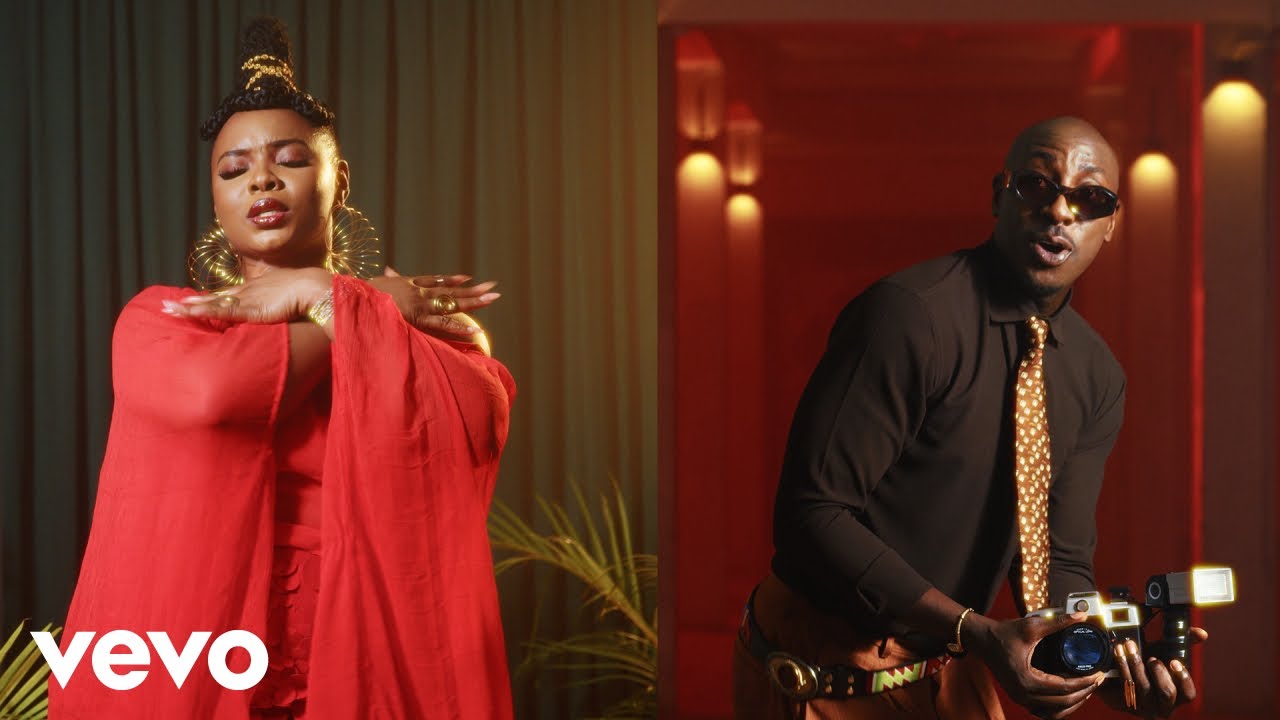 Split-screen music video still from Don't Be Shy featuring Yemi Alade dancing in a sheer red asymmetrical chiffon cape top next to Bien wearing a brown shirt, patterned gold tie, and holding a camera.