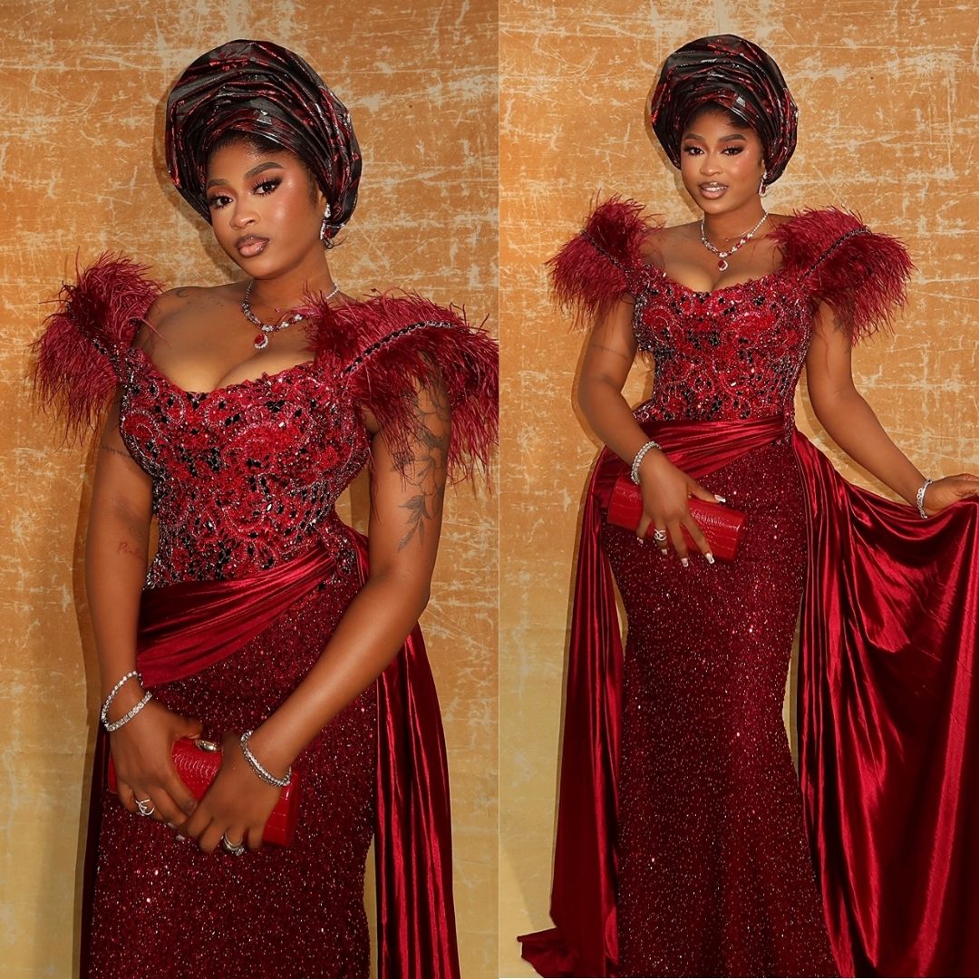 Imisi wearing a crimson sequin corseted gown with feathered shoulder accents, draped waist, matching gele, and a red clutch bag at Peller and Jarvis' traditional wedding.