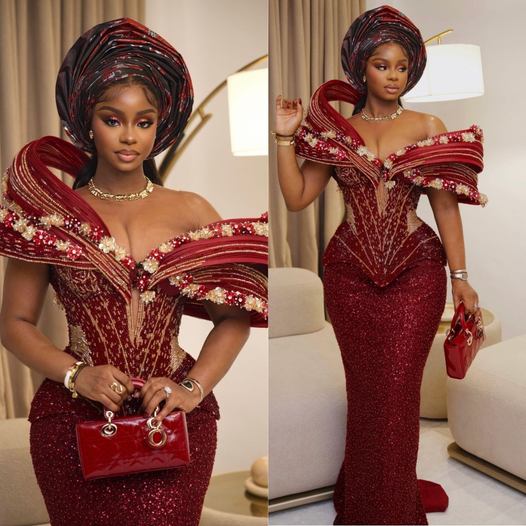 Priscilla Ojo wearing an off-the-shoulder crimson corset gown with sculptured shoulder drapes, gold accents, matching gele, and a red handbag at Peller and Jarvis' traditional wedding.