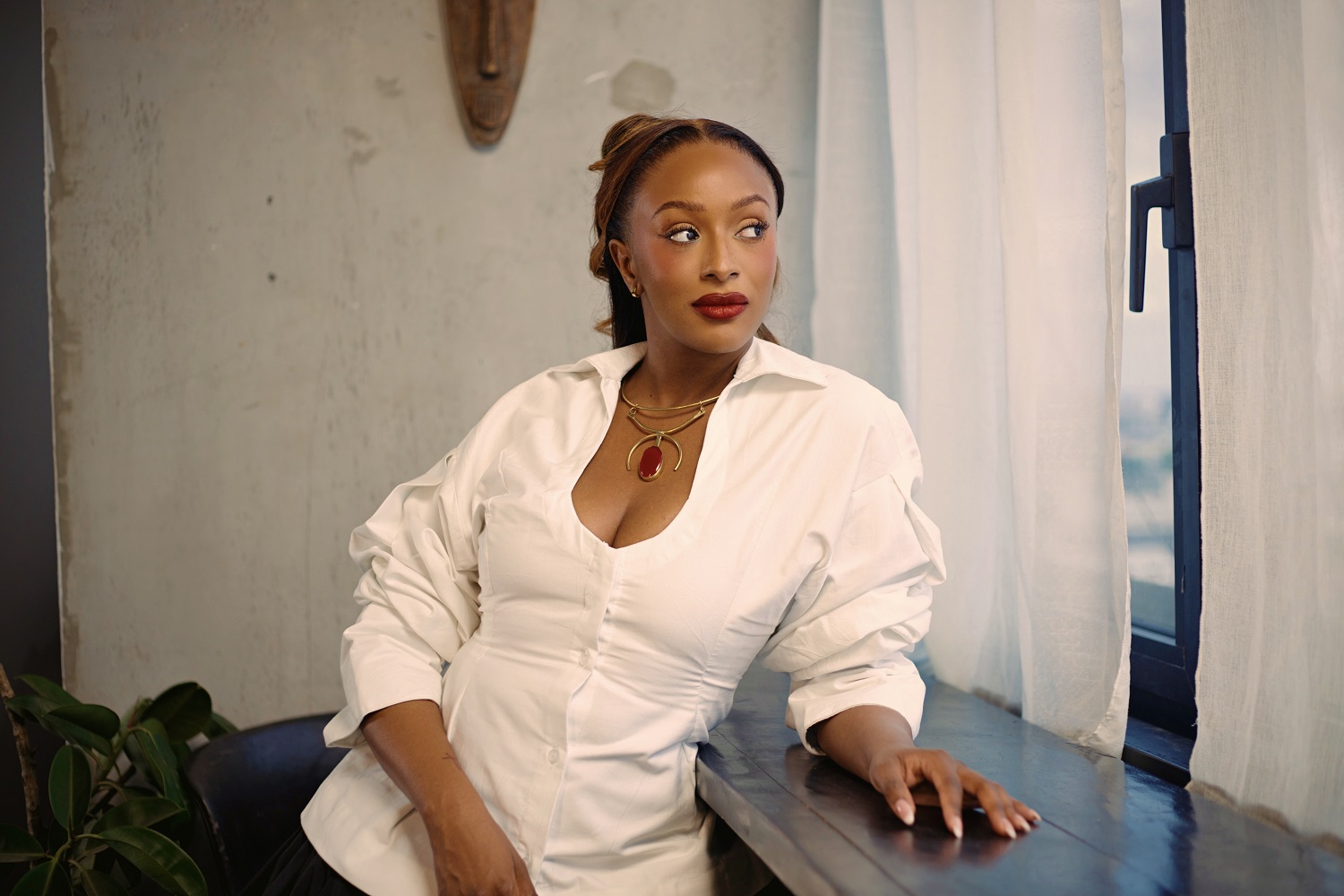 Florence Otedola looking out a window wearing a white collared shirt, reflecting on her philanthropic legacy through the Cuppy Foundation.