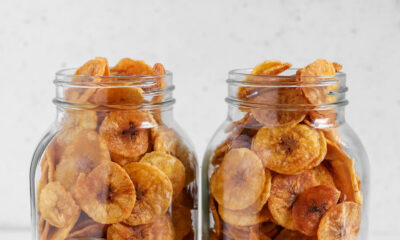 Two clear glass mason jars filled to the brim with thin, golden-brown round plantain chips resting on a table scattered with extra chips.