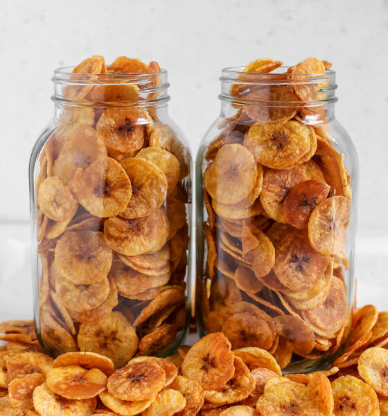 Two clear glass mason jars filled to the brim with thin, golden-brown round plantain chips resting on a table scattered with extra chips.