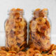 Two clear glass mason jars filled to the brim with thin, golden-brown round plantain chips resting on a table scattered with extra chips.