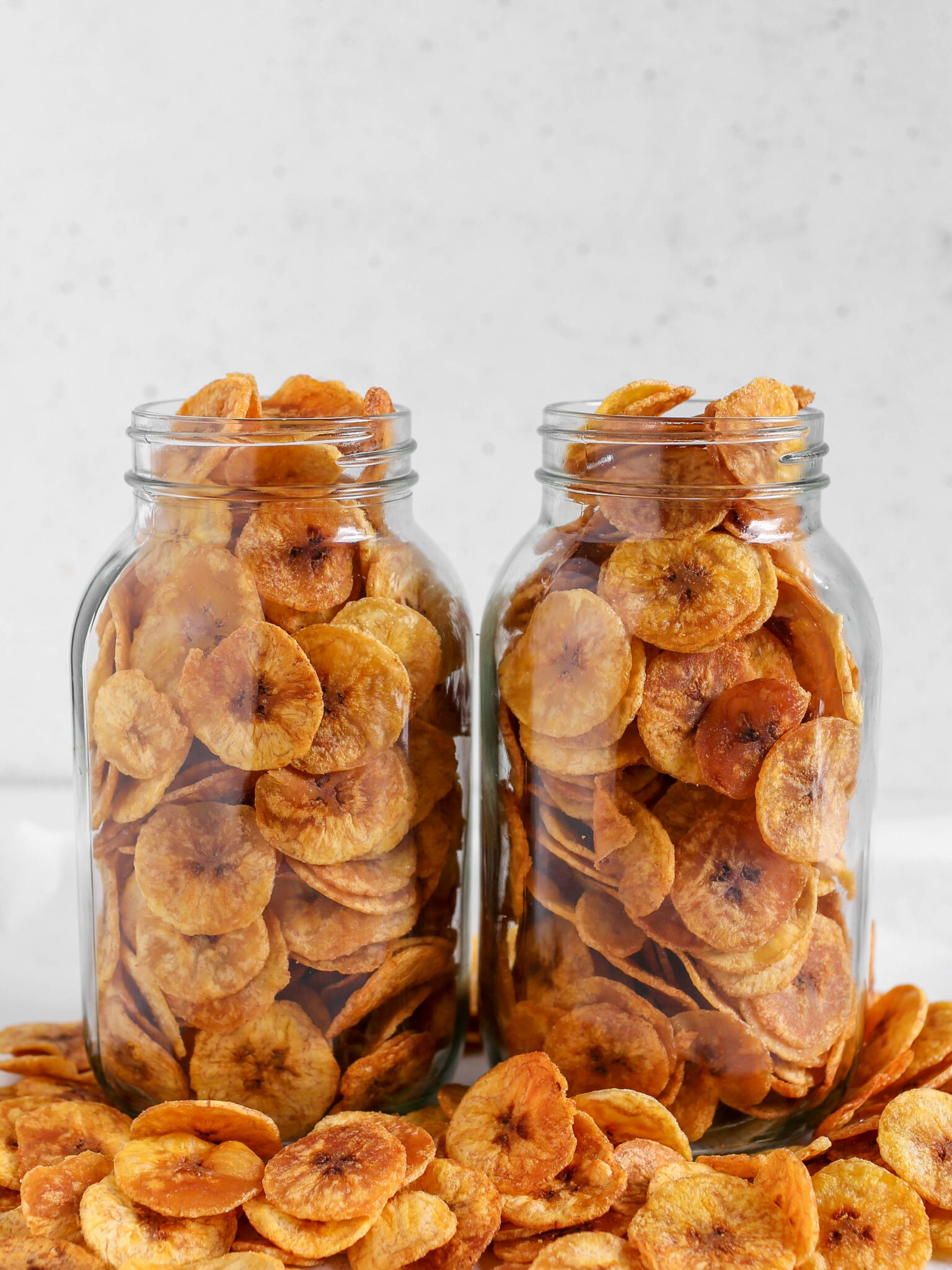 Two clear glass mason jars filled to the brim with thin, golden-brown round plantain chips resting on a table scattered with extra chips.