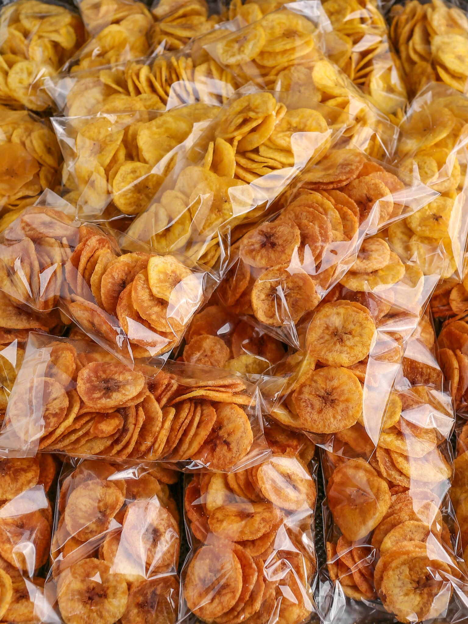 Multiple small clear plastic bags filled with thin, crunchy round plantain chips stacked closely together.