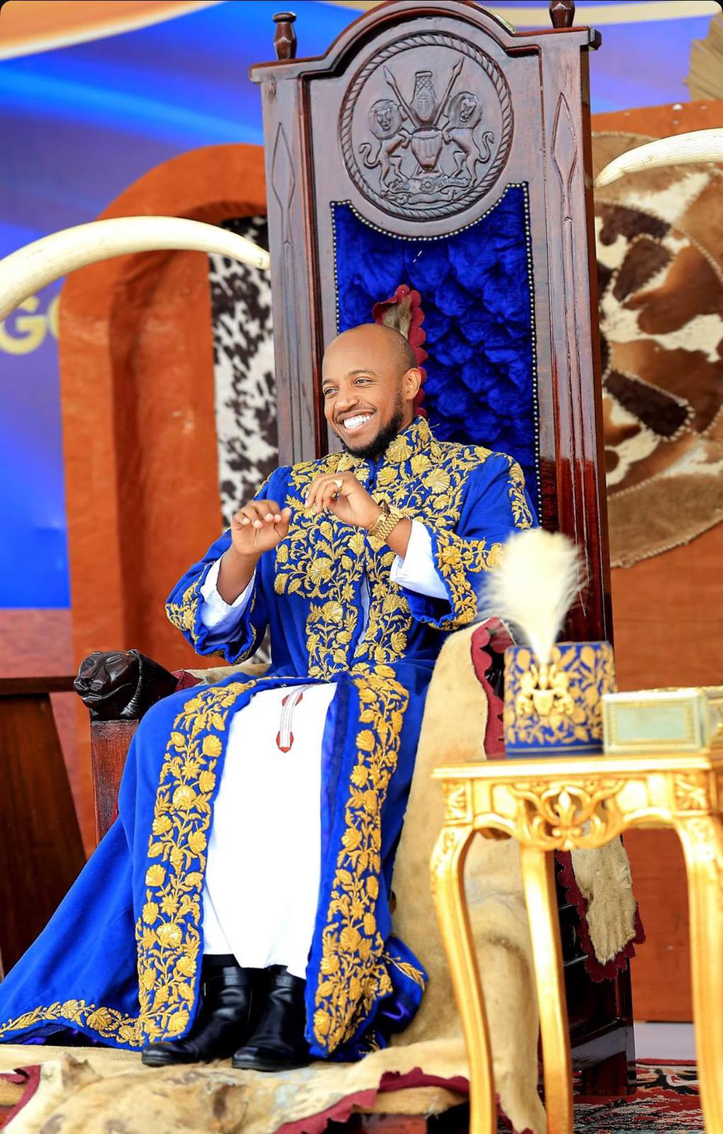 King Oyo Nyimba Kabamba Iguru Rukidi IV smiling on his royal throne in gold-embroidered blue robes during a traditional ceremony.