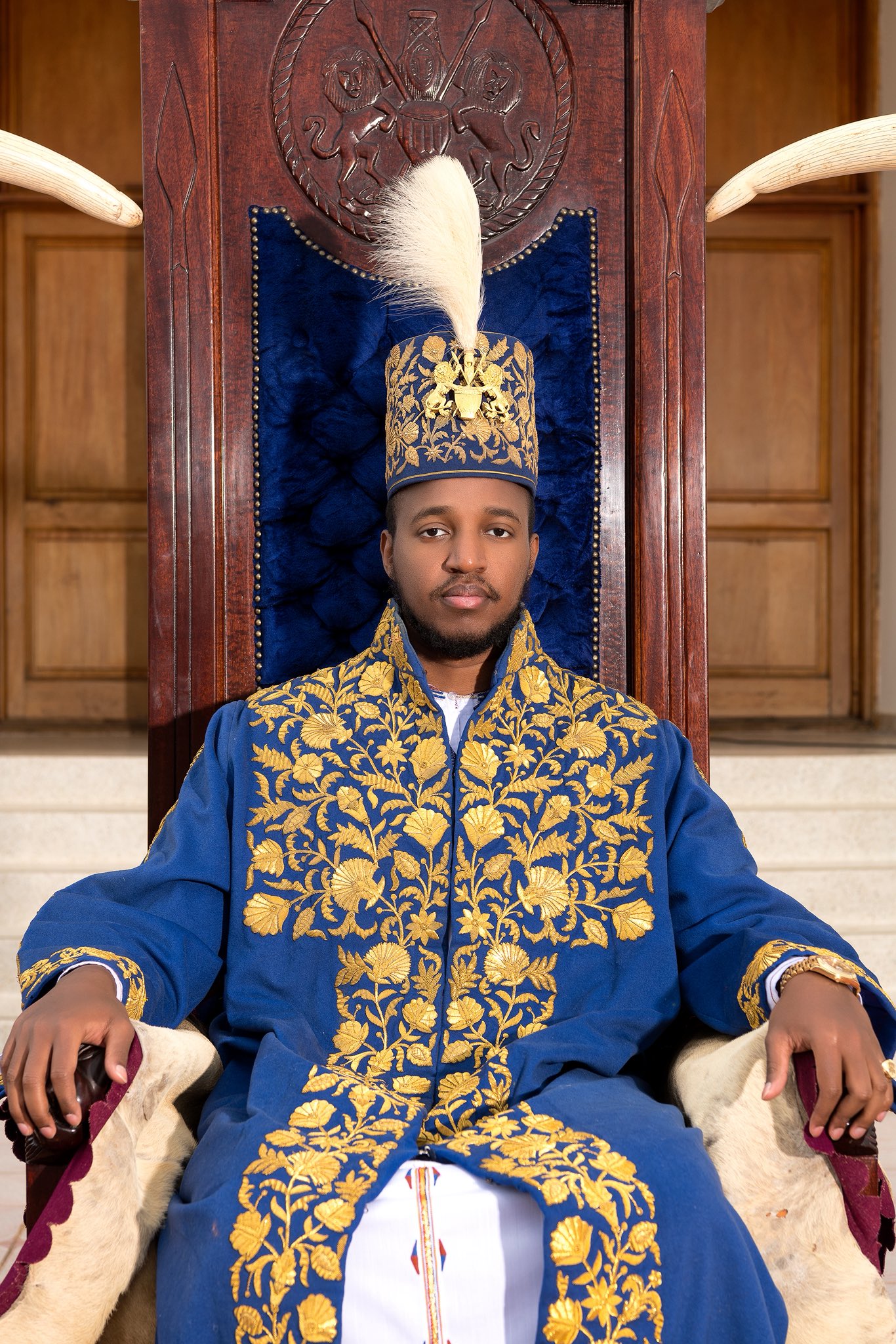 King Oyo Nyimba Kabamba Iguru Rukidi IV seated on the Tooro Kingdom throne in embroidered blue ceremonial robes and a royal feather crown.