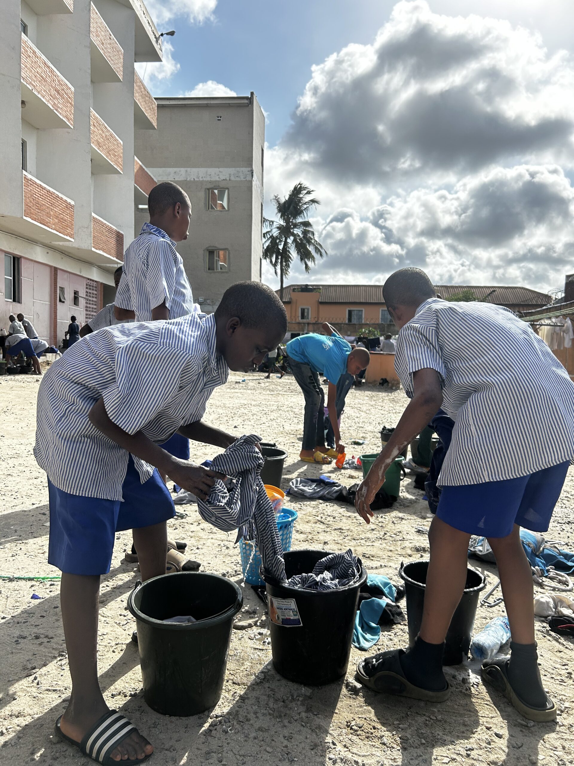 Students in blue-and-white striped uniforms washing clothes in buckets outside the boarding facilities at Let It Shine Academy.