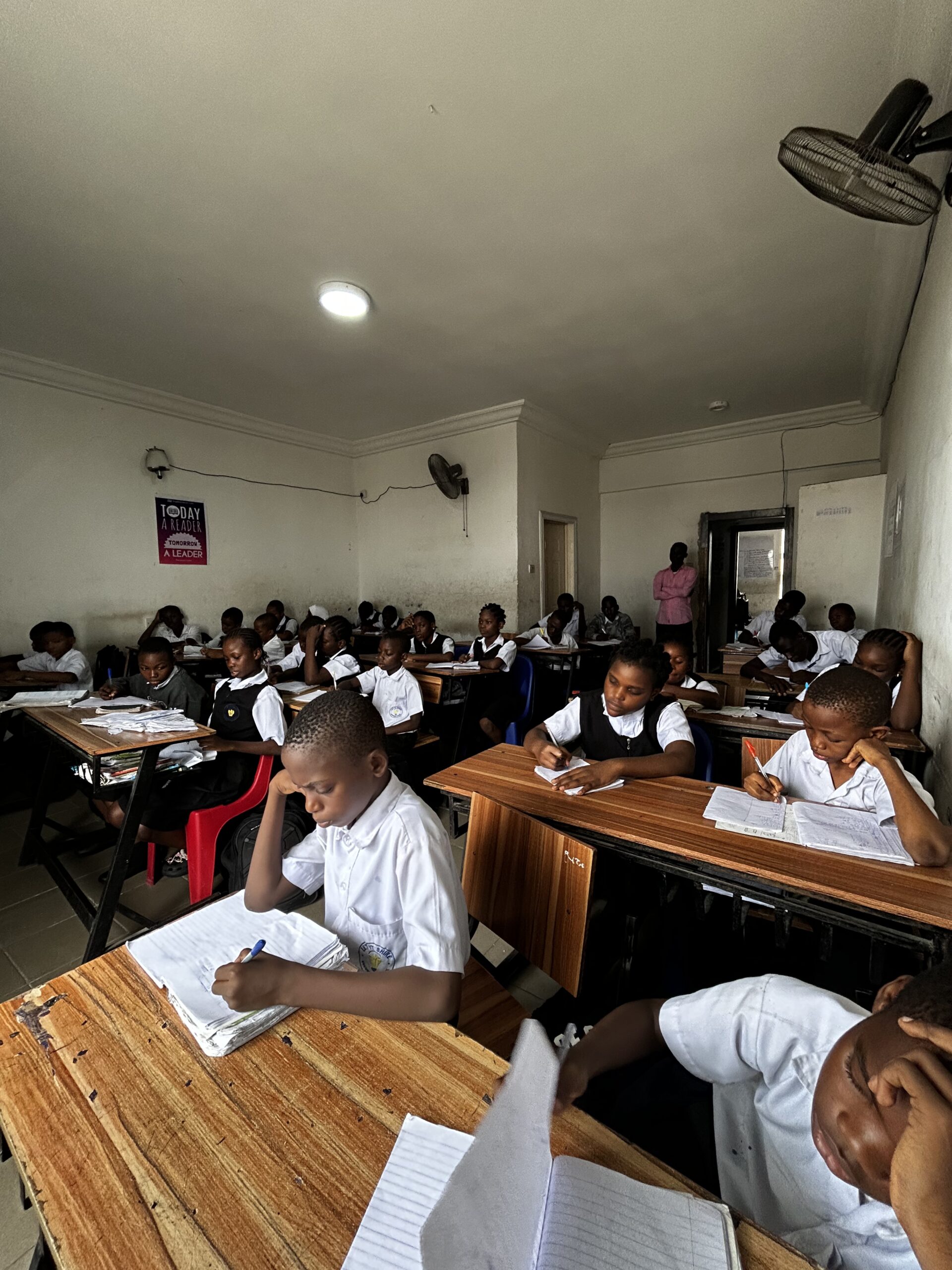 Students in white school uniforms focused on writing in their notebooks inside a classroom at Let It Shine Academy.
