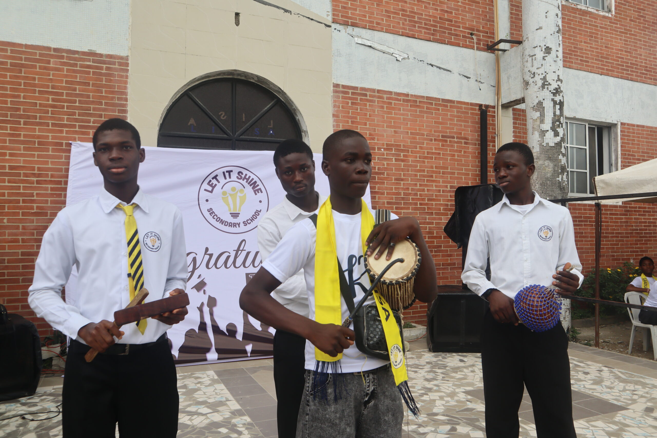 Students in school uniforms performing with traditional African musical instruments including talking drum and shekere at Let It Shine Academy.