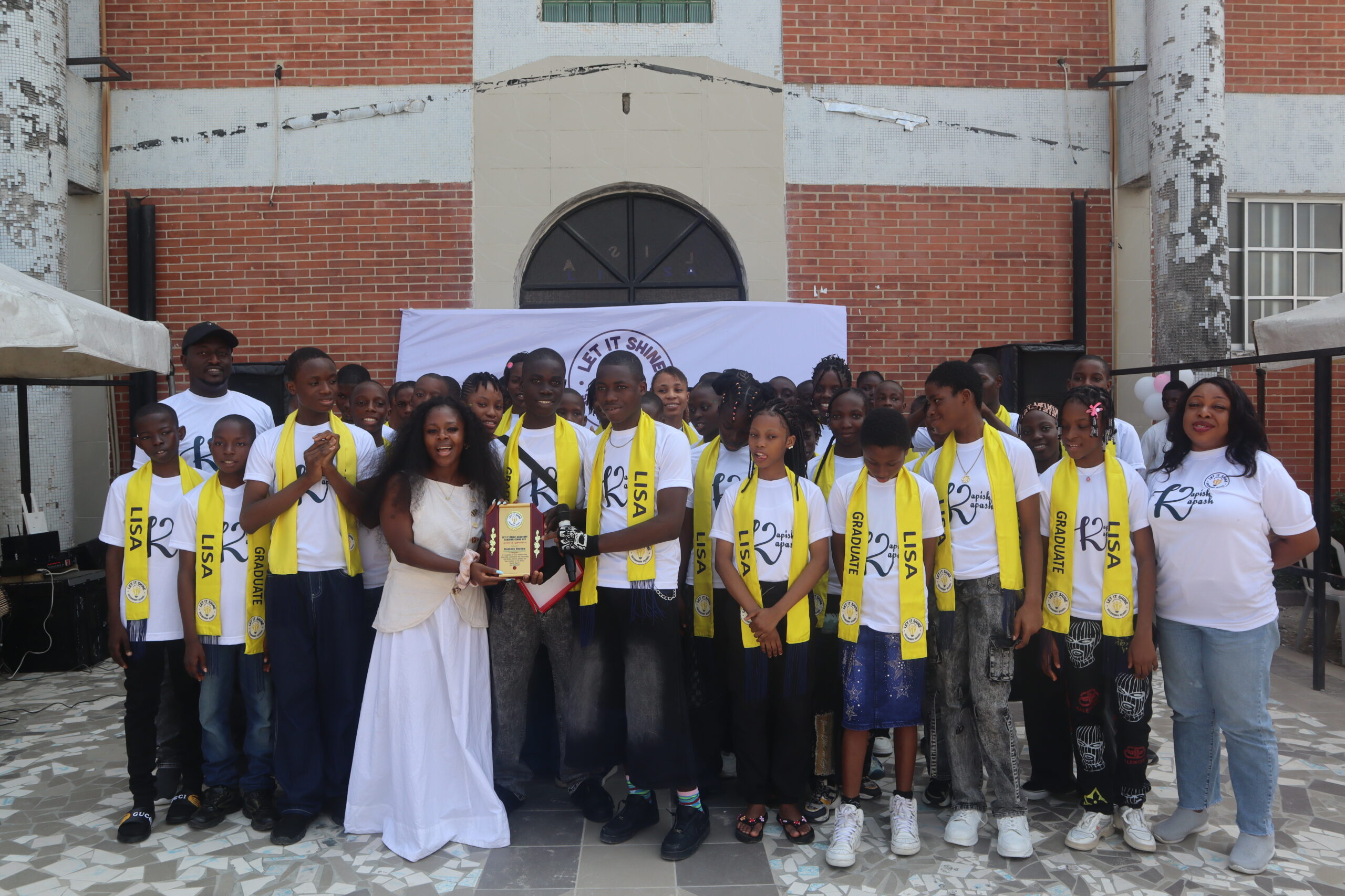 Founder Damilola Feyide posing with graduating students wearing yellow sashes at Let It Shine Academy graduation.