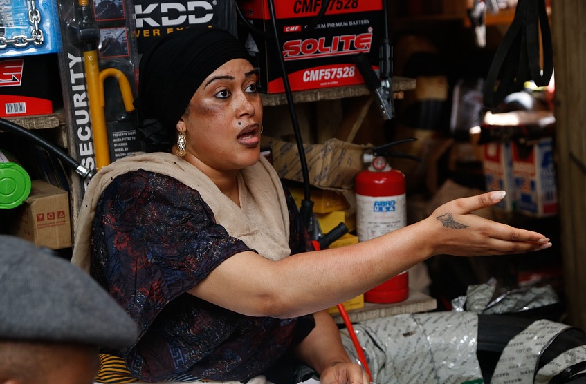 Adunni Ade, in character as Iya Mulika, wears a headwrap and speaks emphatically in an auto parts shop during the filming of Tokunbo.