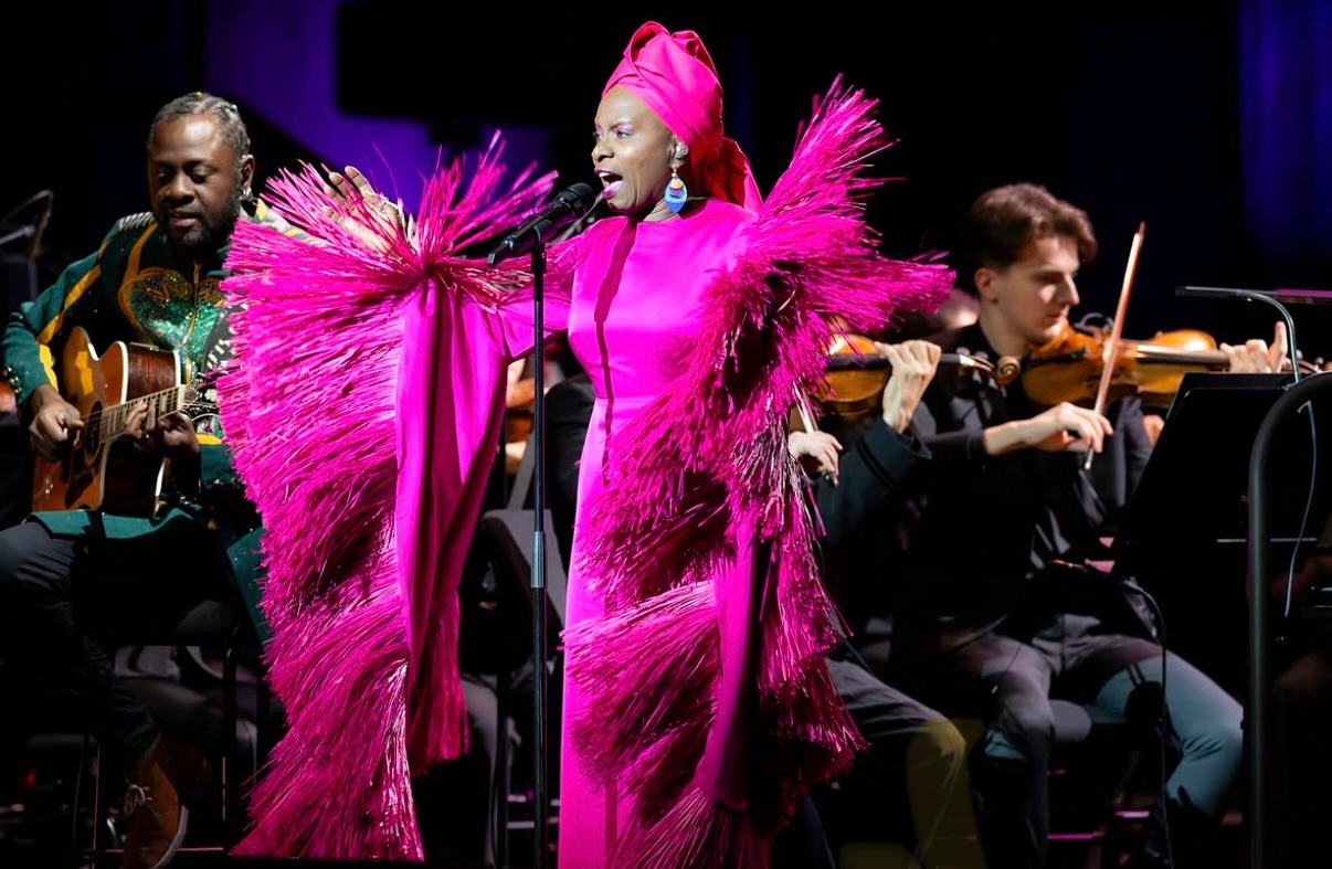 Angélique Kidjo singing onstage in a bright pink fringed gown and matching headwrap with the Philharmonie de Paris orchestra in 2025.