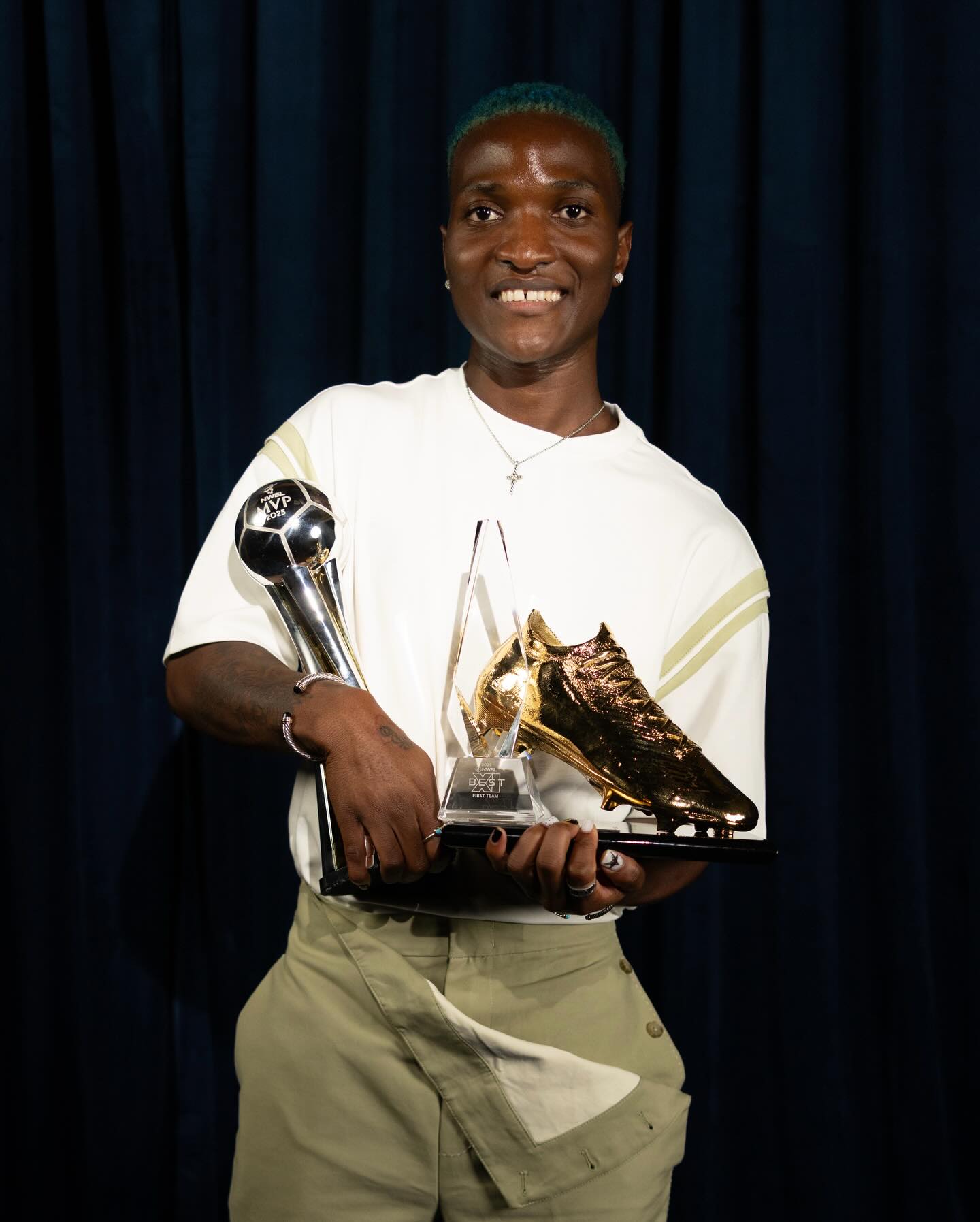 Temwa Chawinga wearing a white top and green trousers while holding her NWSL MVP and Golden Boot trophies.