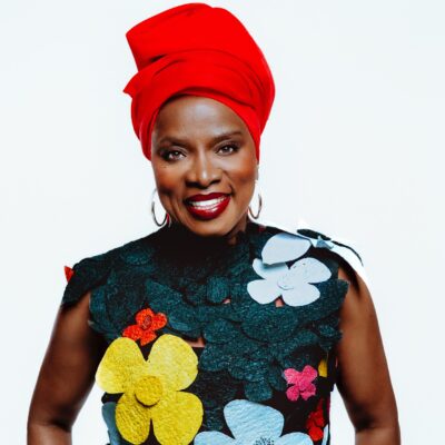 Studio portrait of Benin-born singer Angélique Kidjo smiling in a floral appliqué sleeveless top and a vibrant red headwrap.