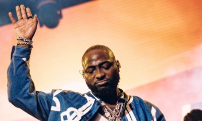 Afrobeats artist Davido performing live on stage at Coachella 2026 with one hand raised, wearing a denim jacket and diamond chain.