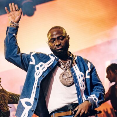 Afrobeats artist Davido performing live on stage at Coachella 2026 with one hand raised, wearing a denim jacket and diamond chain.