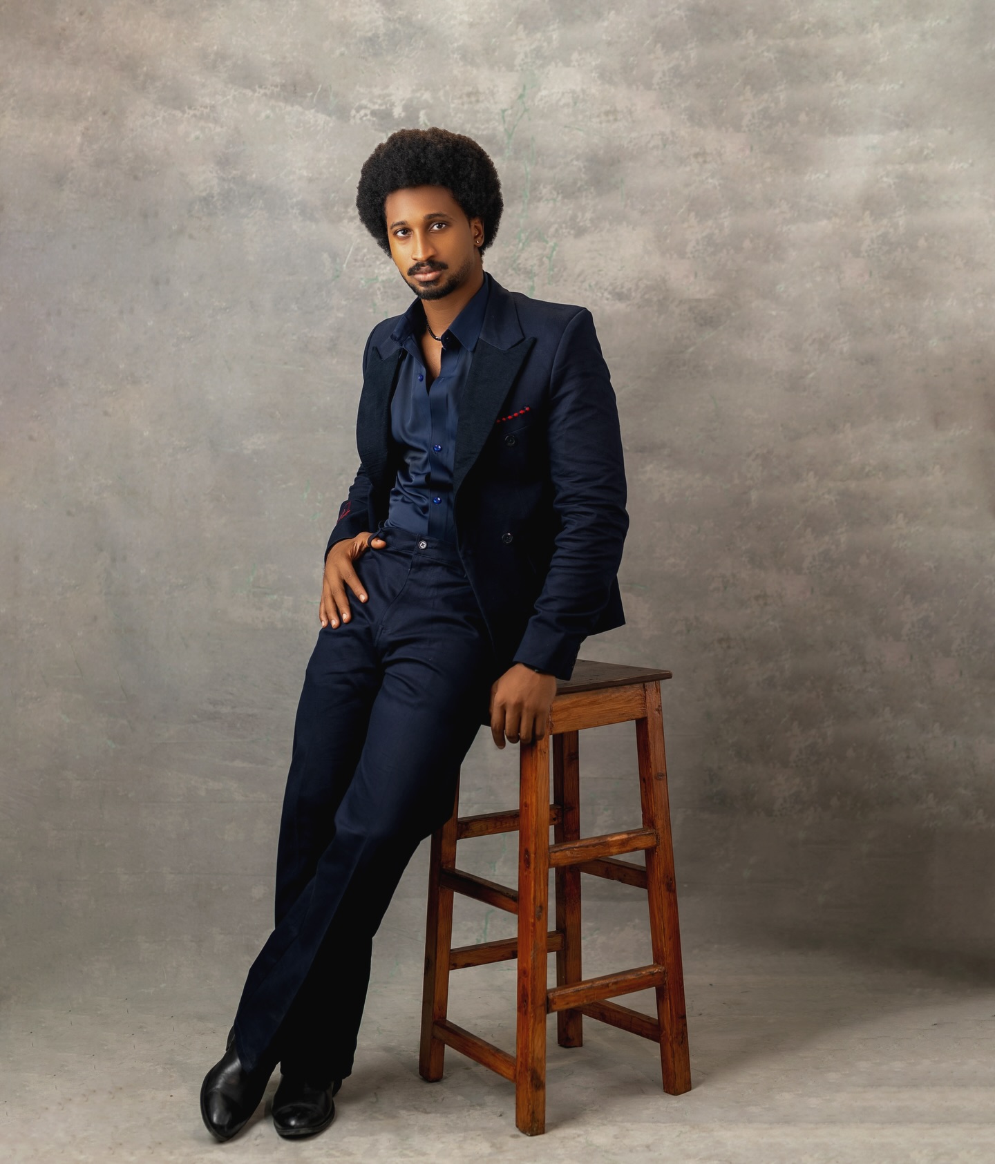 Chimsom Chuka leaning against a wooden stool in a navy double-breasted suit and black boots.