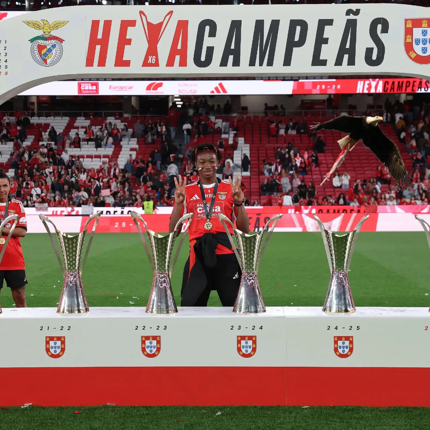 Michaely Bihina gesturing victory behind four trophies on the pitch following SL Benfica's Portuguese title celebrations.