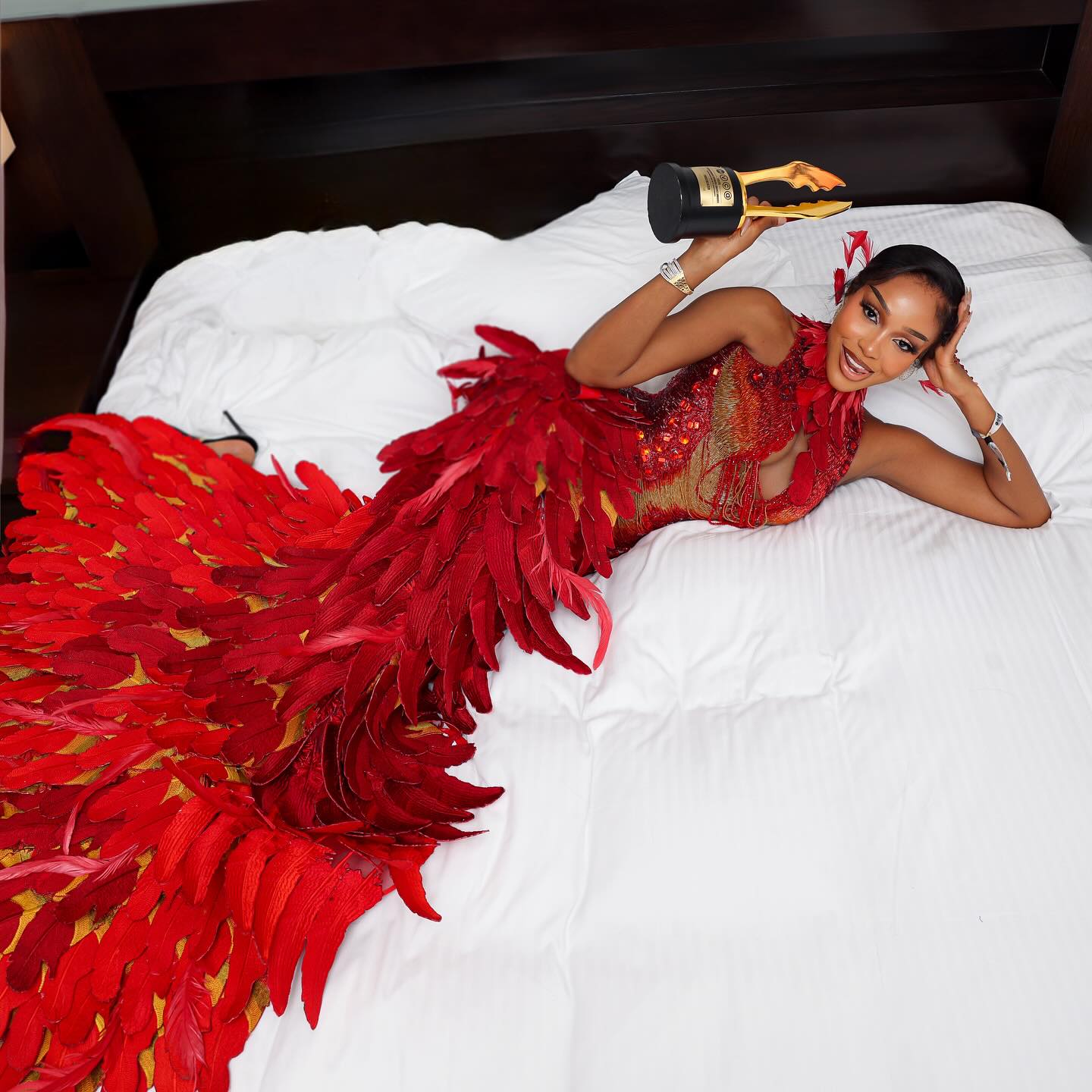 Uche Montana holding her Trailblazer Award trophy in a red feathered dress.