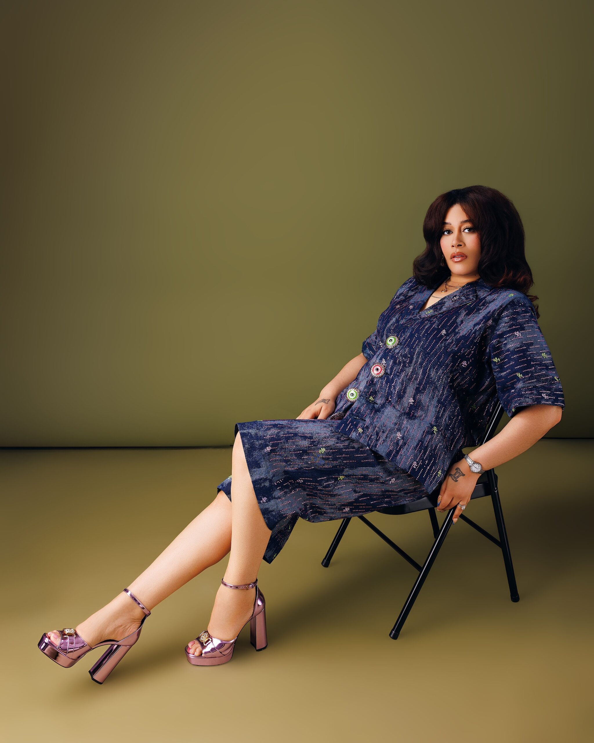Adunni Ade wearing a navy patterned jacket, resting her arm on a chair while looking into the camera.