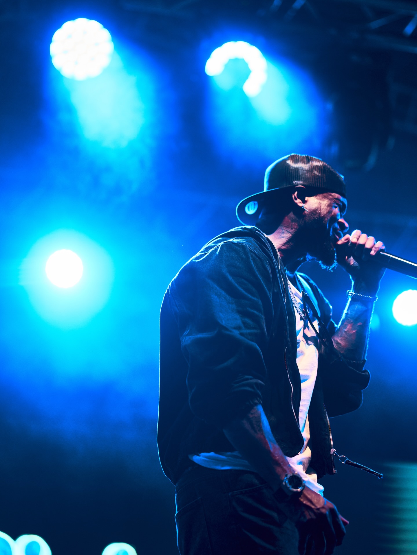 Mr P singing into a microphone on stage lit by blue concert lights, wearing a dark jacket over a white shirt, a backward cap, and wrist accessories.
