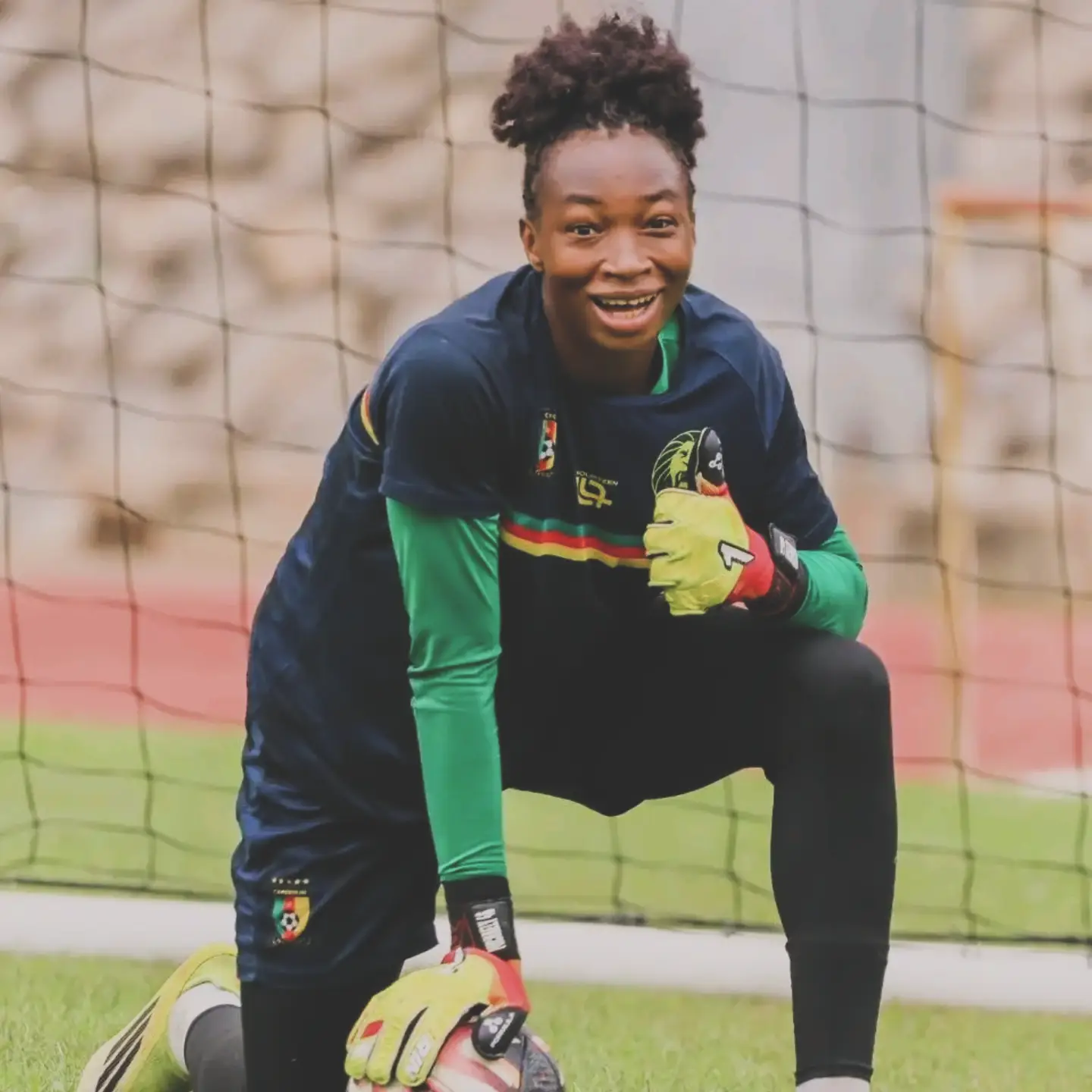 Cameroonian goalkeeper Michaely Bihina kneeling on the pitch in her national team training gear while holding a football.