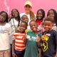 Shakira wearing a yellow cap standing behind eight members of Uganda's Triplets Ghetto Kids smiling while holding ice cream cups in front of a pink wall.