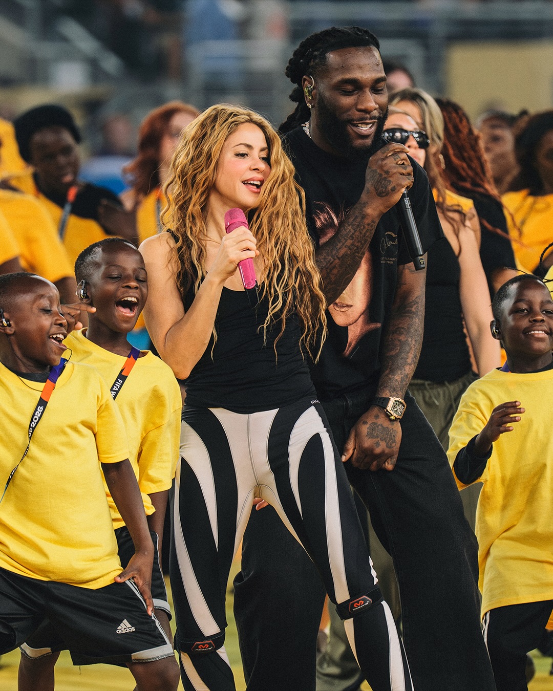 Burna Boy and Shakira performing live on stage surrounded by children in yellow shirts.