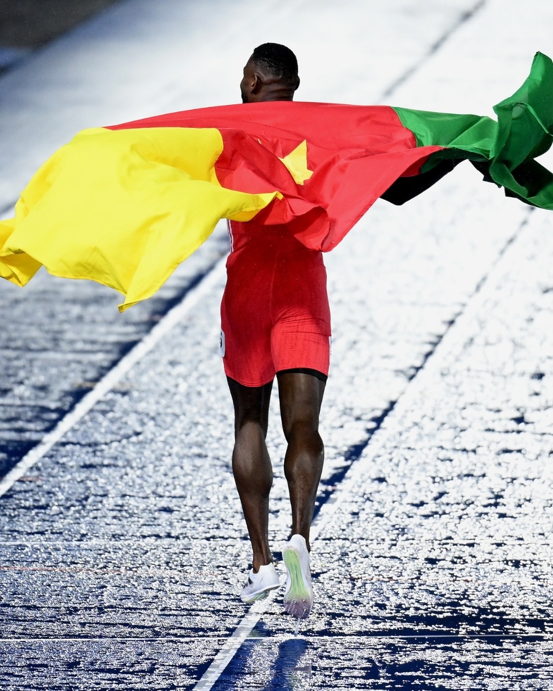 Emmanuel Eseme carrying the green, red, and yellow Cameroonian national flag on the athletics track following his 100m gold medal win.
