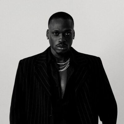 Black and white portrait of Nigerian Afrobeats artist Fireboy DML wearing a dark pinstripe suit jacket and layered silver chains ahead of his CLAAT single release.