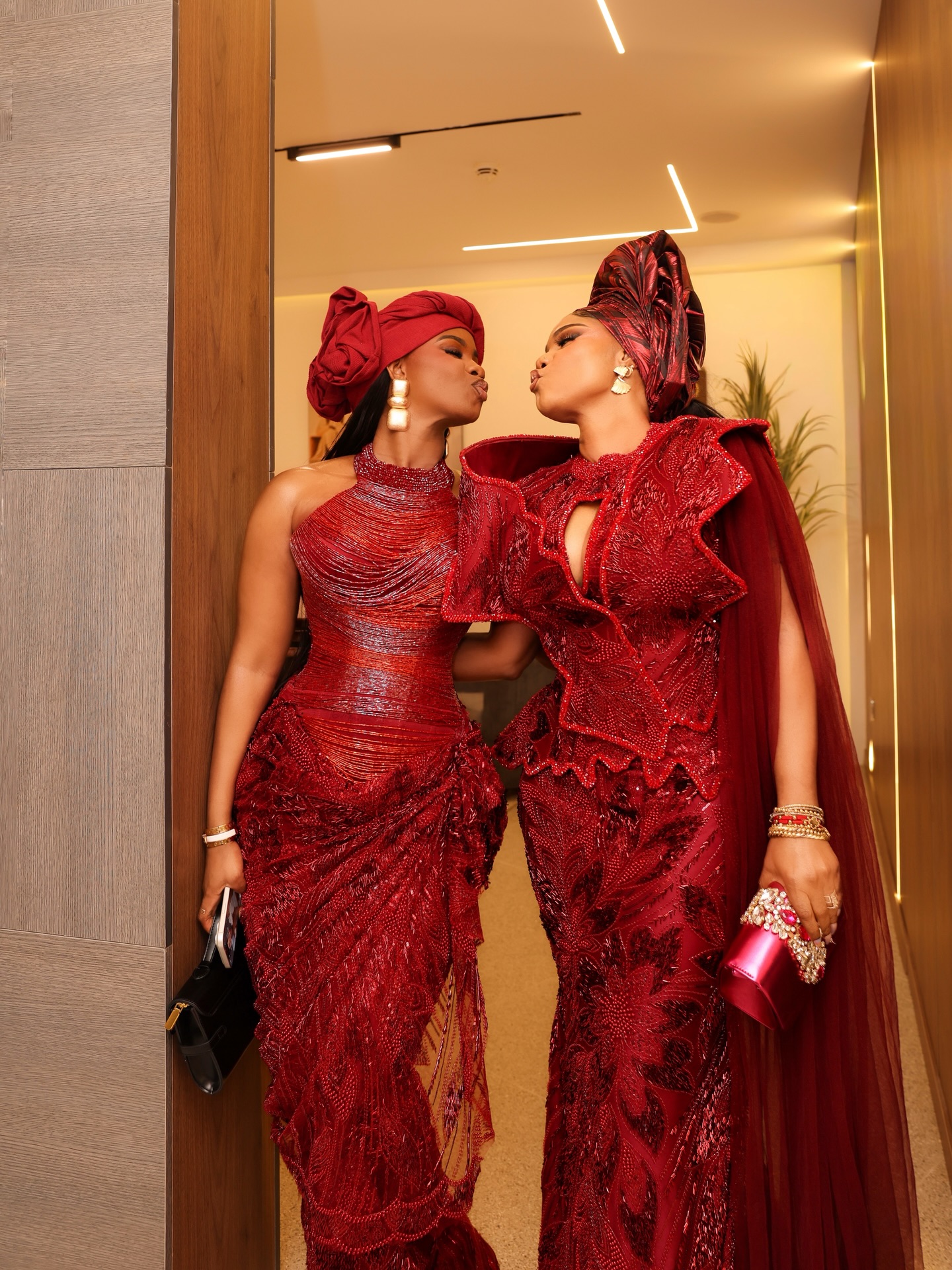 Iyabo Ojo and her daughter Priscilla Ojo sharing an air kiss in custom crimson red embellished lace gowns and coordinated Gele headwraps.