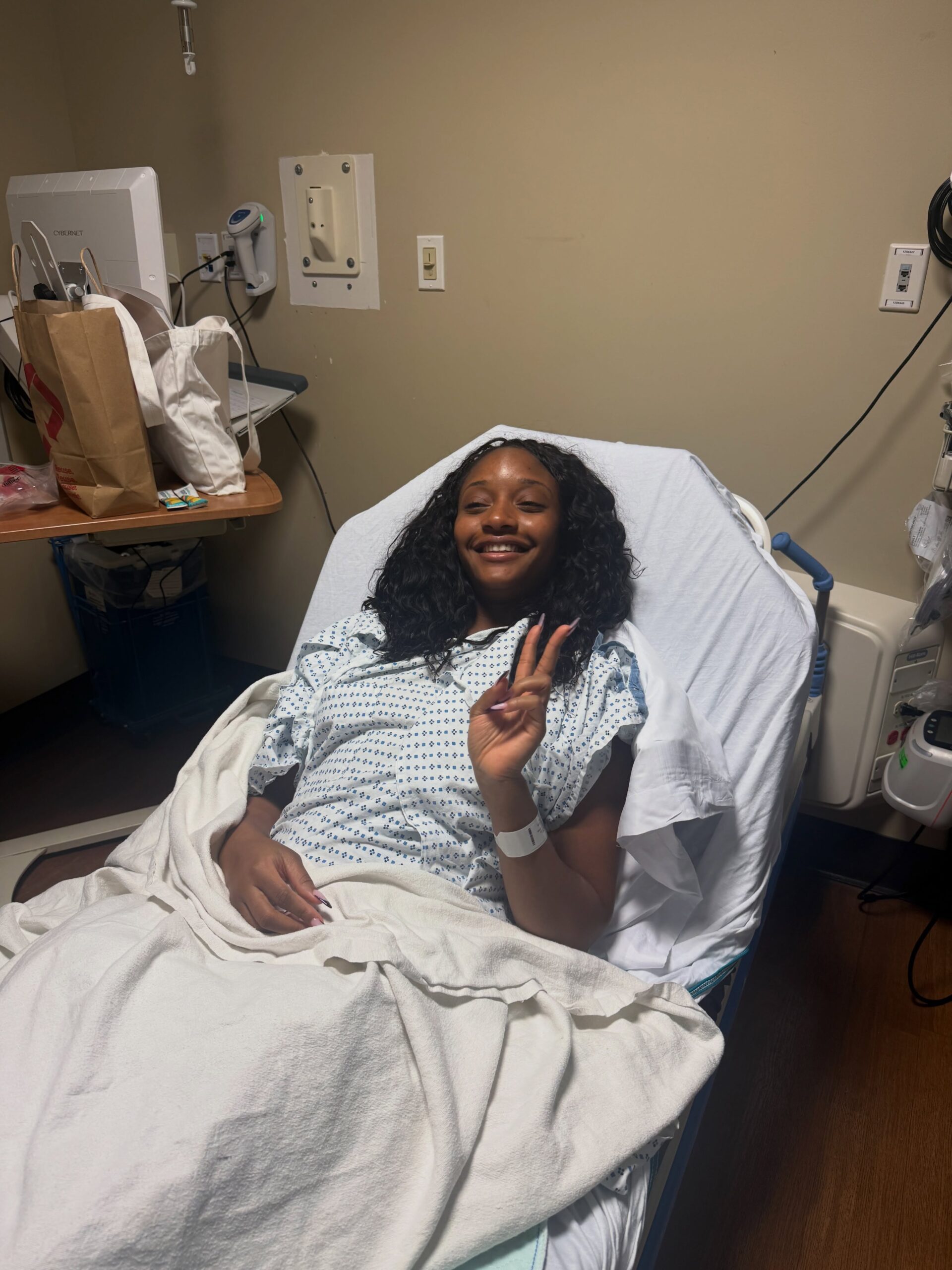 Ayra Starr smiling and giving a peace sign from a hospital bed wearing a medical gown following major surgery.