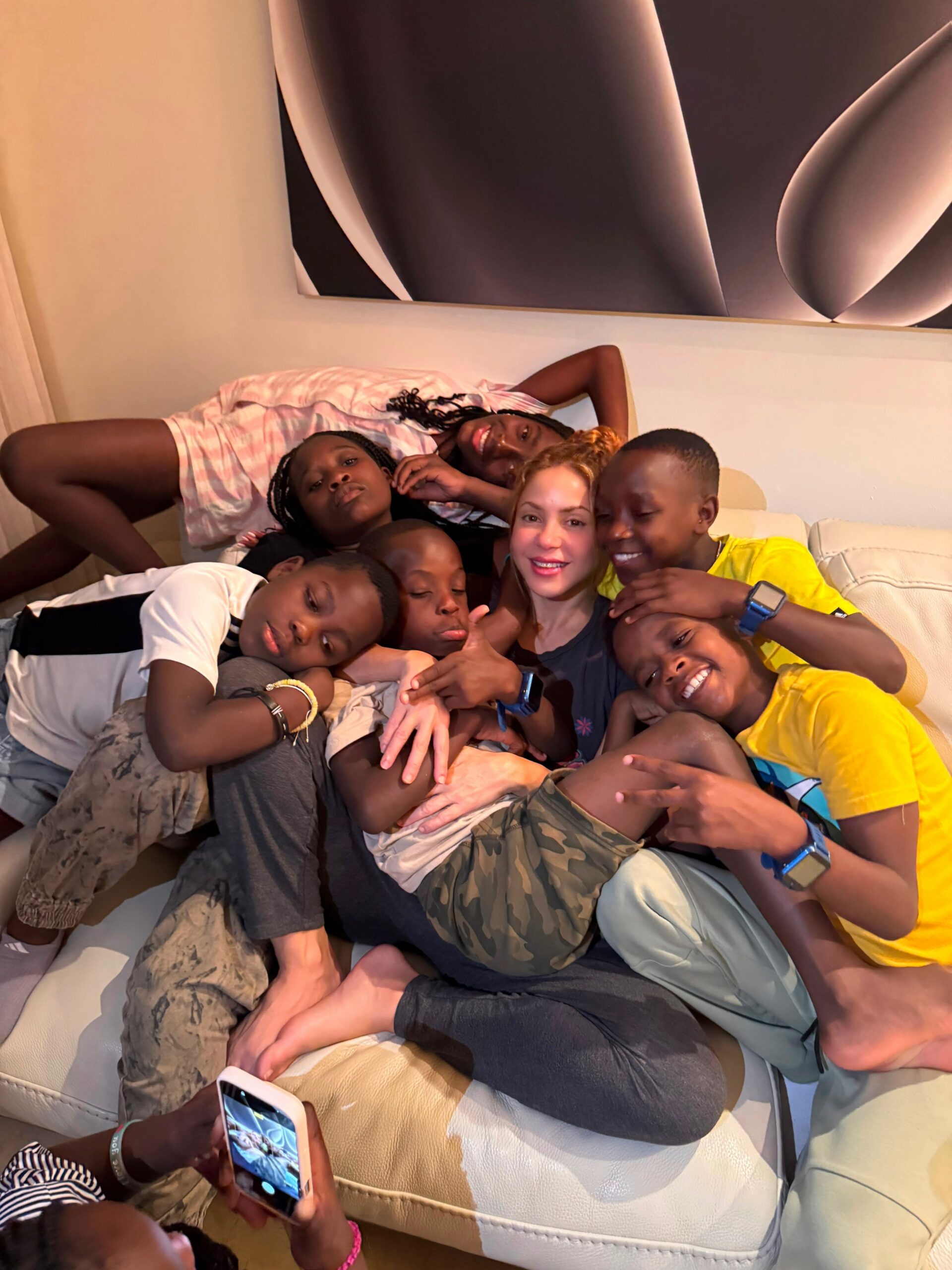 Shakira sitting surrounded by members of the Triplets Ghetto Kids resting together on a white leather sofa indoors.