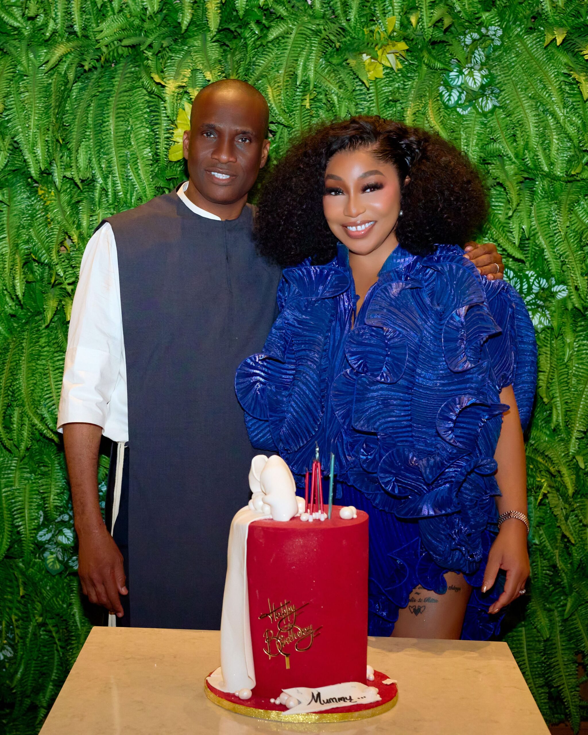 Nollywood actress Rita Dominic and her husband Fidelis Anosike standing beside a red birthday cake decorated with white ribbon icing during her birthday dinner.