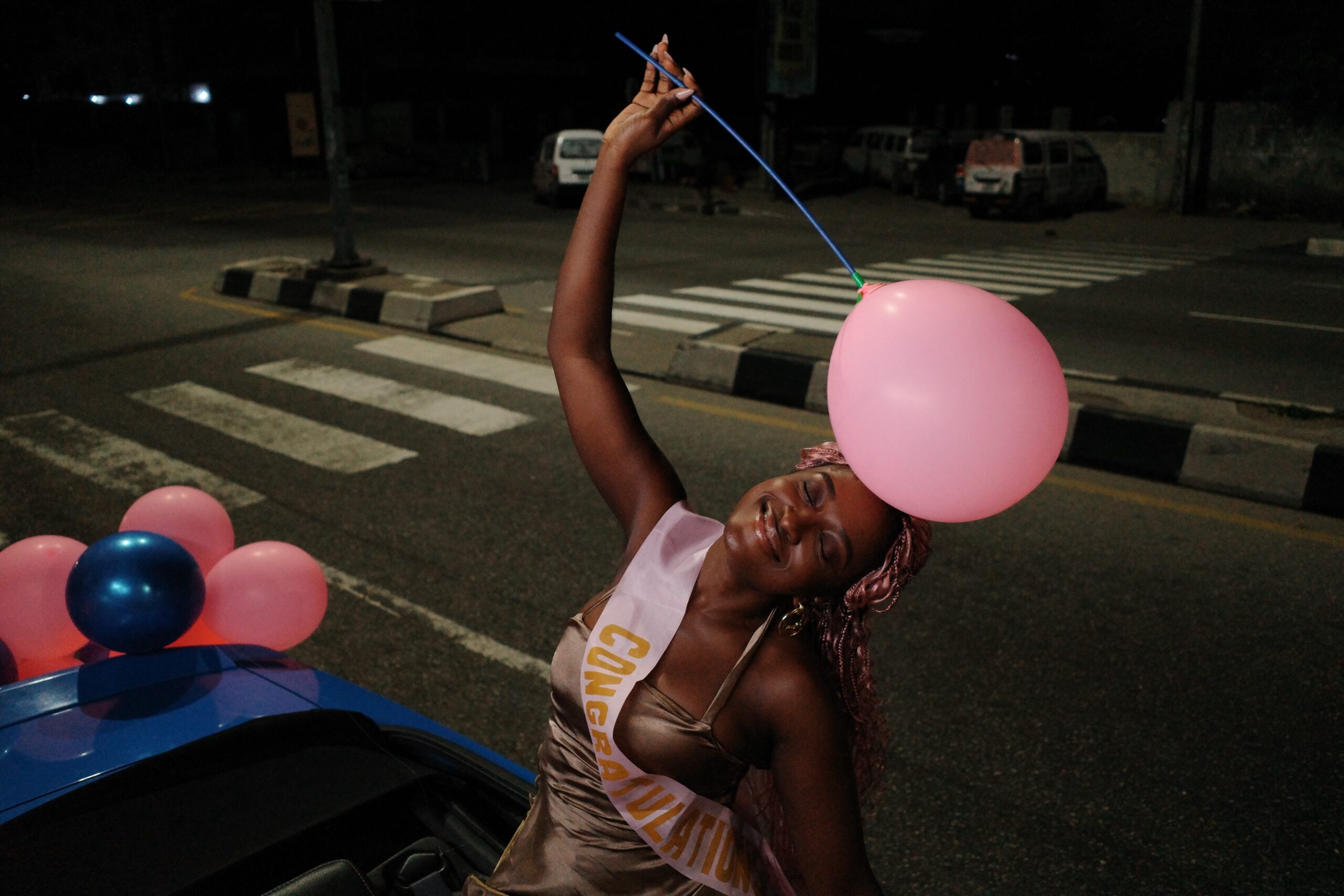 Genoveva Umeh wearing a congratulations sash holding a pink balloon outdoors in a convertible vehicle scene from Siraam.