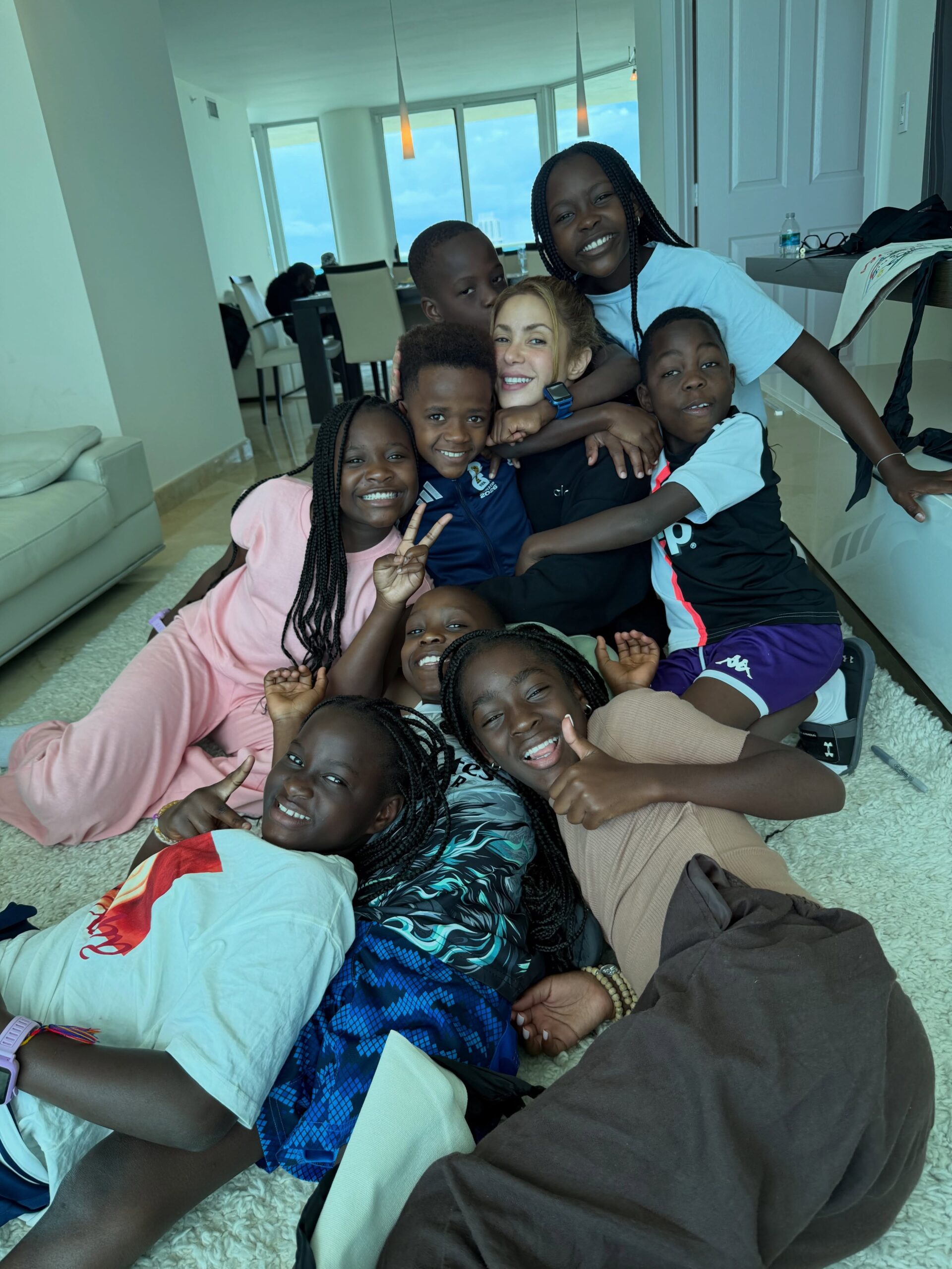 Shakira sitting on a rug surrounded by all eight members of the Triplets Ghetto Kids smiling and giving thumbs-up gestures.