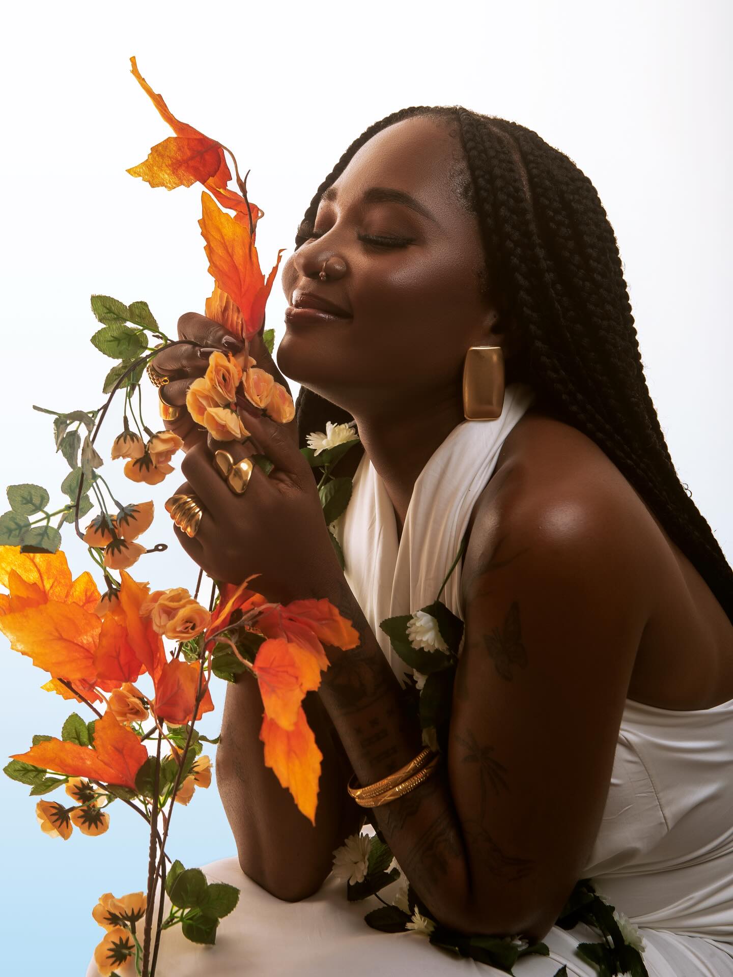 Goddessa posing with autumn foliage wearing a white halter outfit, long box braids, and gold jewelry.