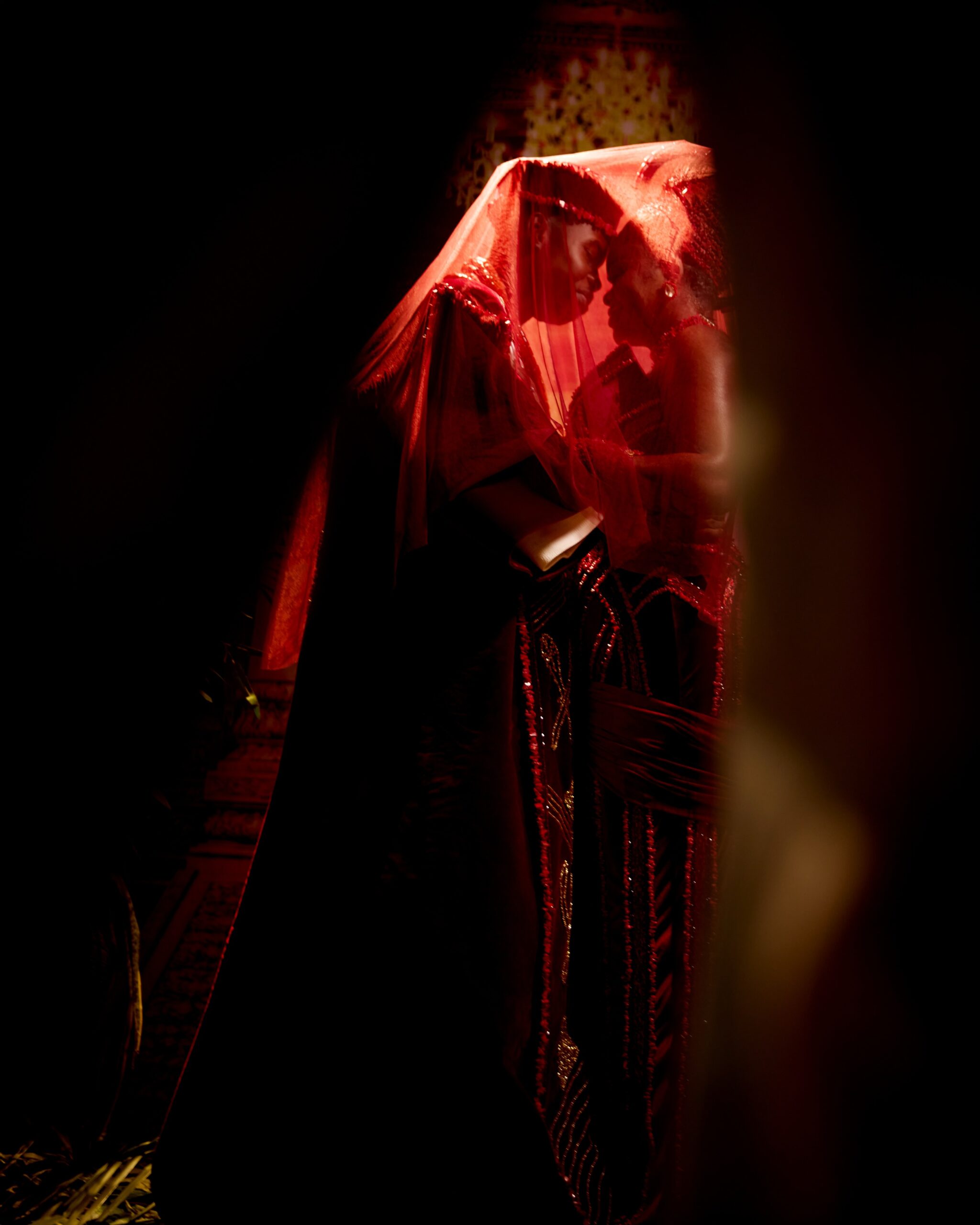 Peller and Jarvis standing together under a sheer red bridal veil during their traditional Edo wedding ceremony portrait.