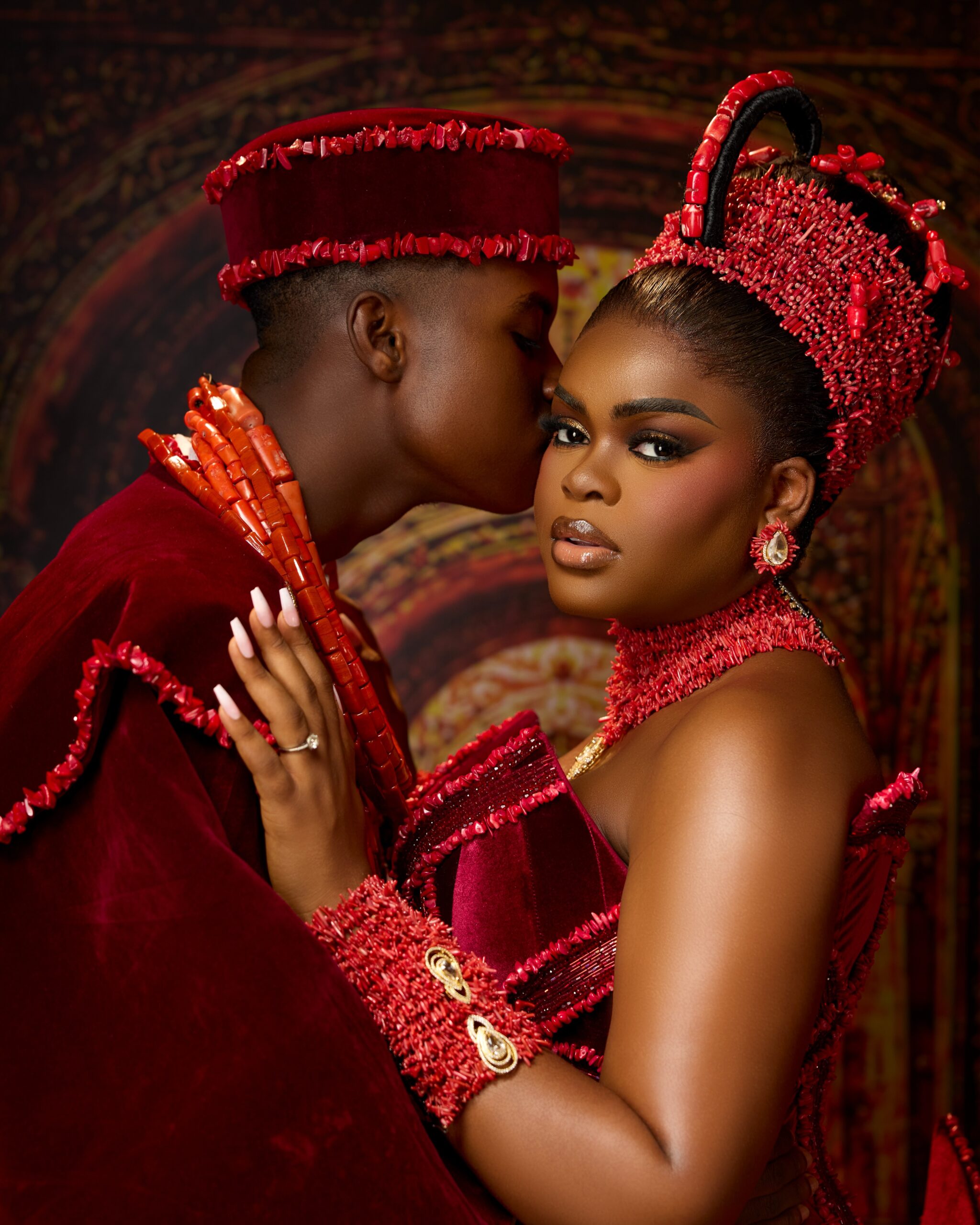 Peller kissing Jarvis on the cheek during their traditional Edo wedding celebration, showing her Okuku beaded crown and coral choker necklace.