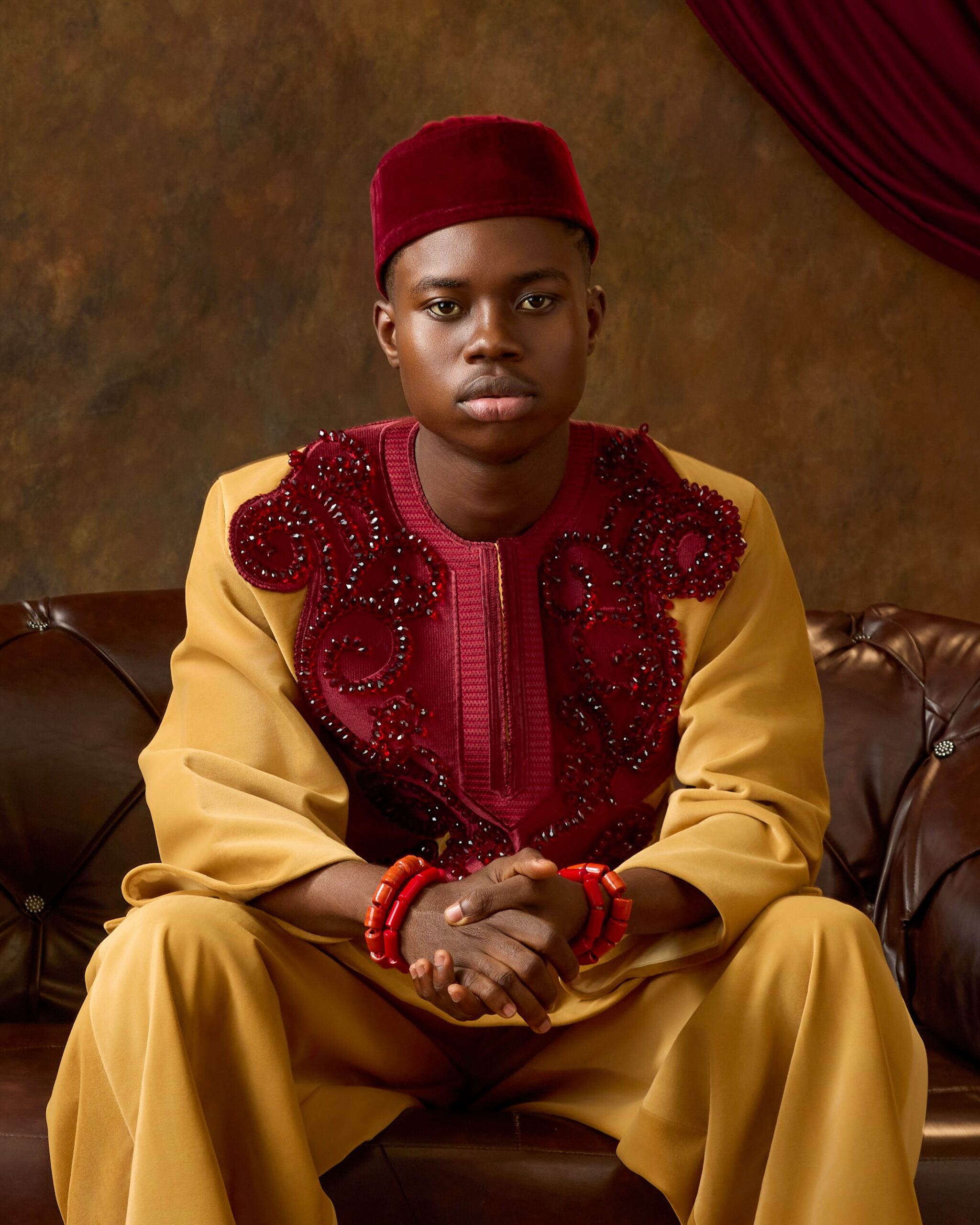 Peller seated on a leather sofa wearing a mustard-gold traditional tunic with a burgundy velvet embroidered bib and Okpu Agwu cap.