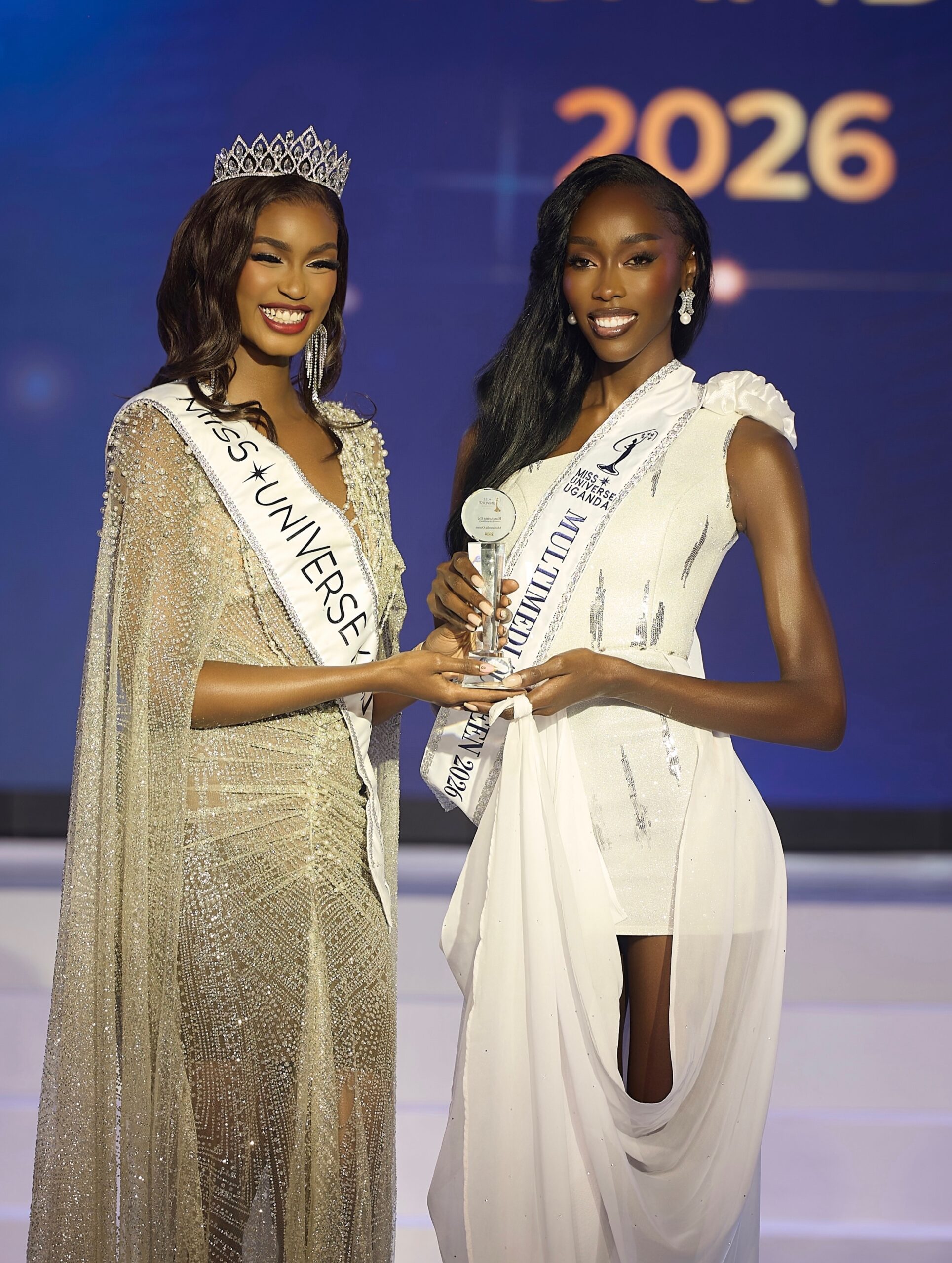 Miss Universe Kenya 2026 Jean Ojiro presenting the Multimedia Queen award trophy to Miss Universe Uganda 2026 Mitchelle Daka on stage.