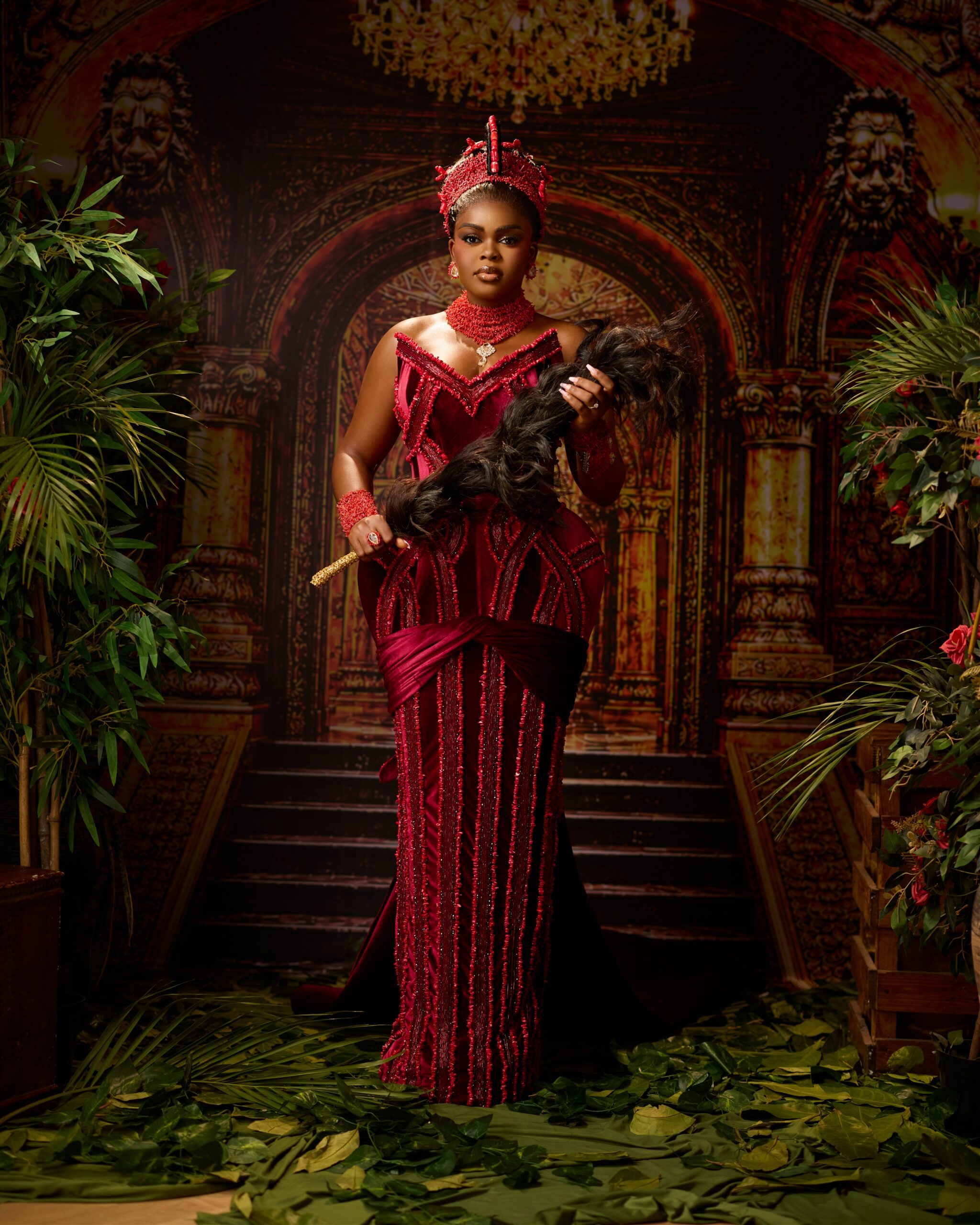 Jarvis posing in a crimson velvet Veekee James gown, Okuku beaded crown, coral choker, and holding a black horsetail fly whisk for her Edo traditional wedding.