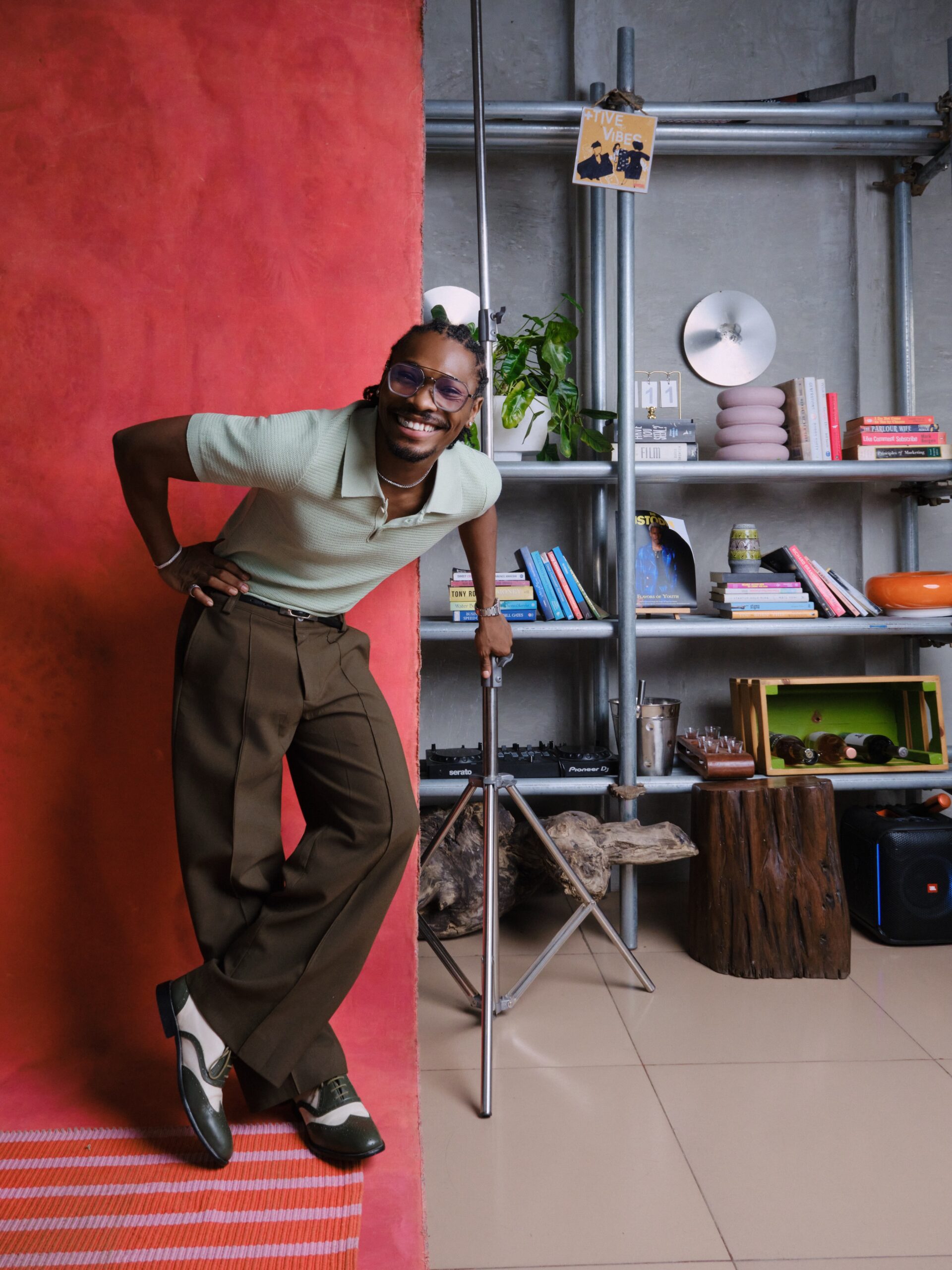 Mike Afolarin wearing a pastel mint green knit polo, pleated olive brown trousers, clear-frame glasses, and spectator shoes.