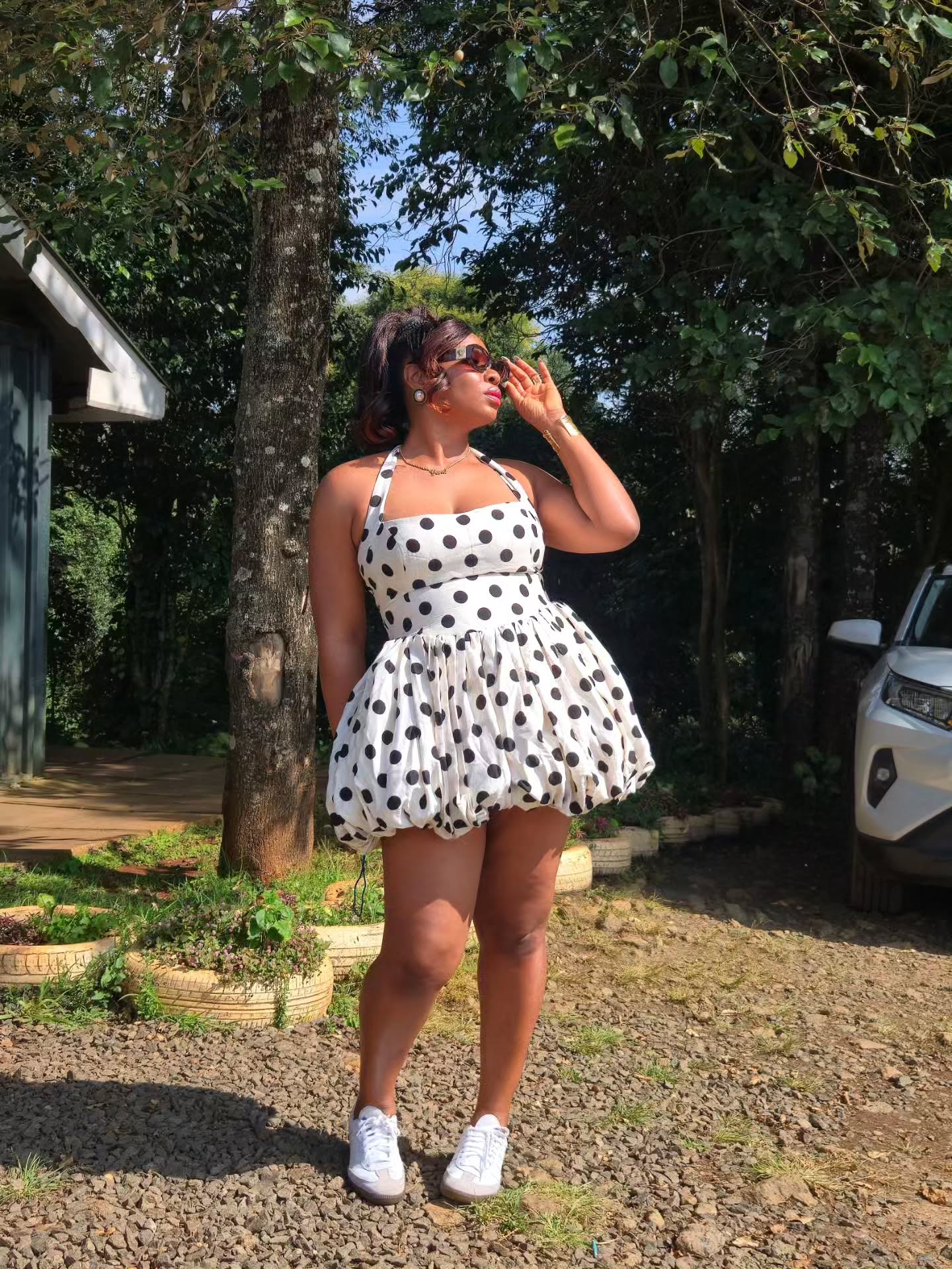 Yemi Alade posing outdoors in a white polka dot halterneck bubble dress with sunglasses and white low-top trainers.