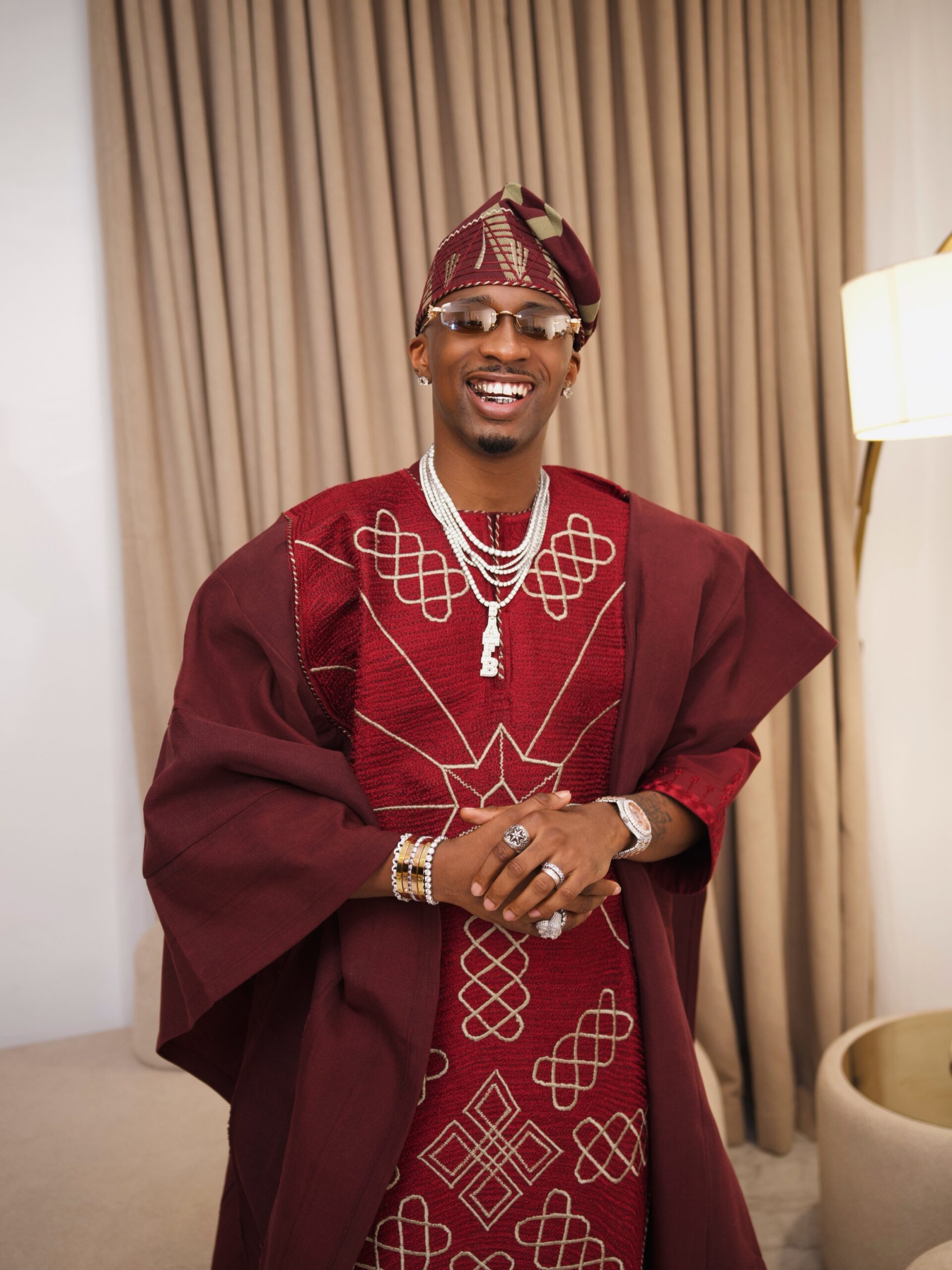 Juma Jux wearing a burgundy embroidered Agbada by Deji and Kola with matching Fila cap and diamond chains.